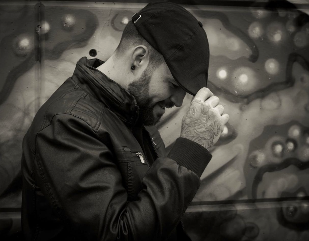 Black and white profile of Nathan Henry. He is wearing a black leather jacket and black newsboy cap, and he's smiling with his face tipped downward, hand closest to the screen adjusting the brim of his cap.