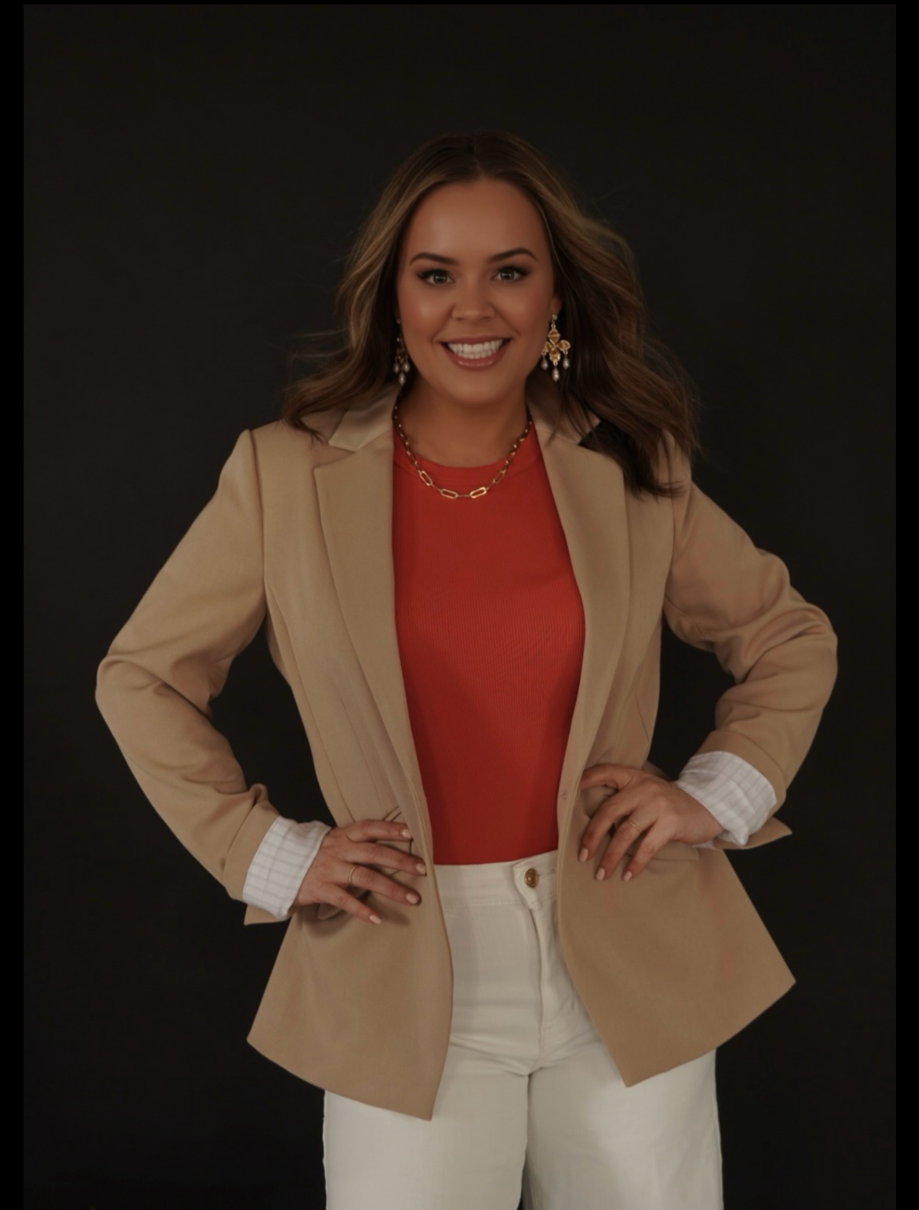 Lisa Campbell smiling with hands on hips, wearing a beige blazer over a red top, white pants, earrings, and jewelry, standing against a black background.