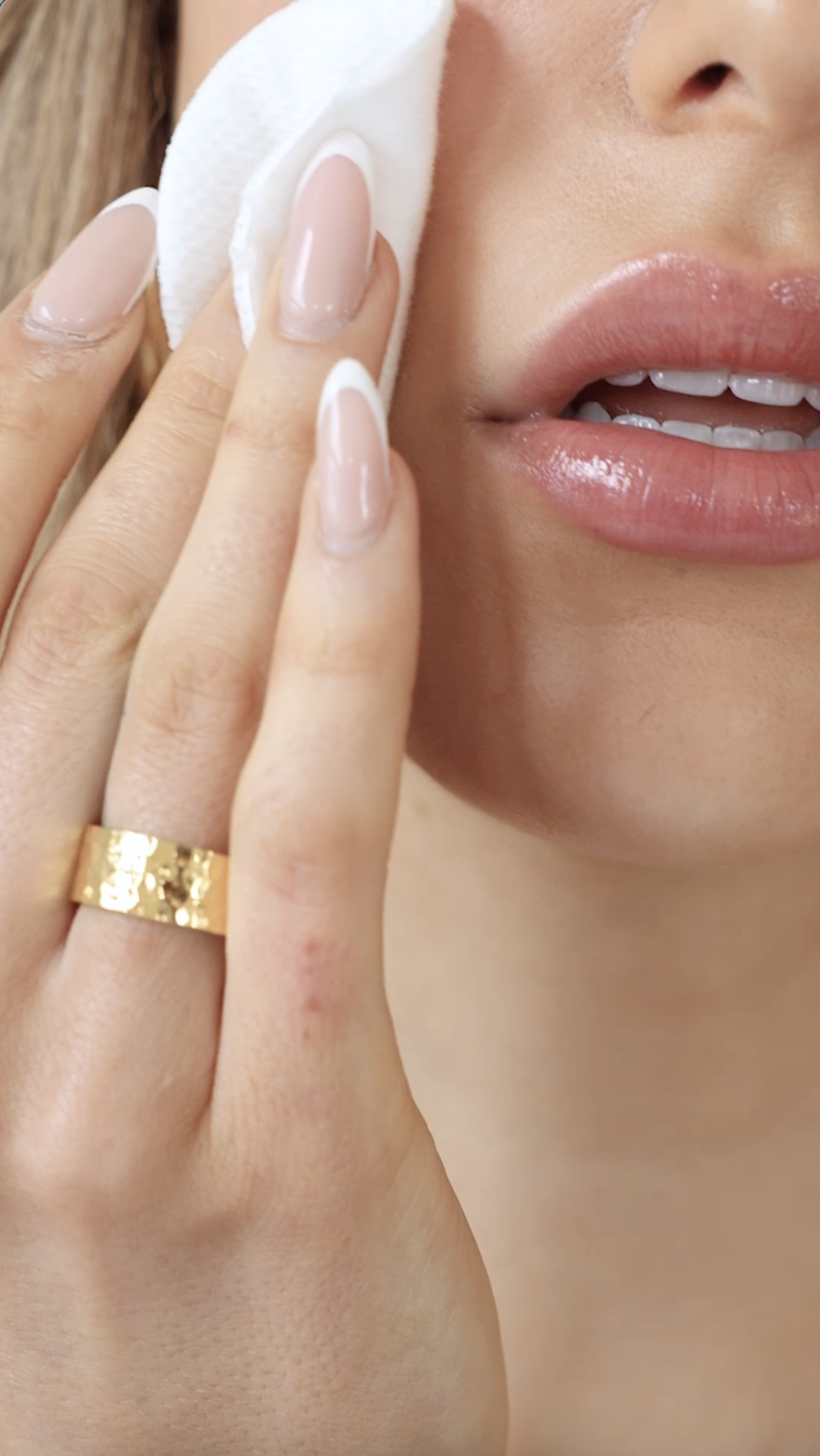 Close-up of a woman's face showing lips, part of her nose, and her hand with neatly manicured nails and a gold ring, holding a cotton pad near her cheek.