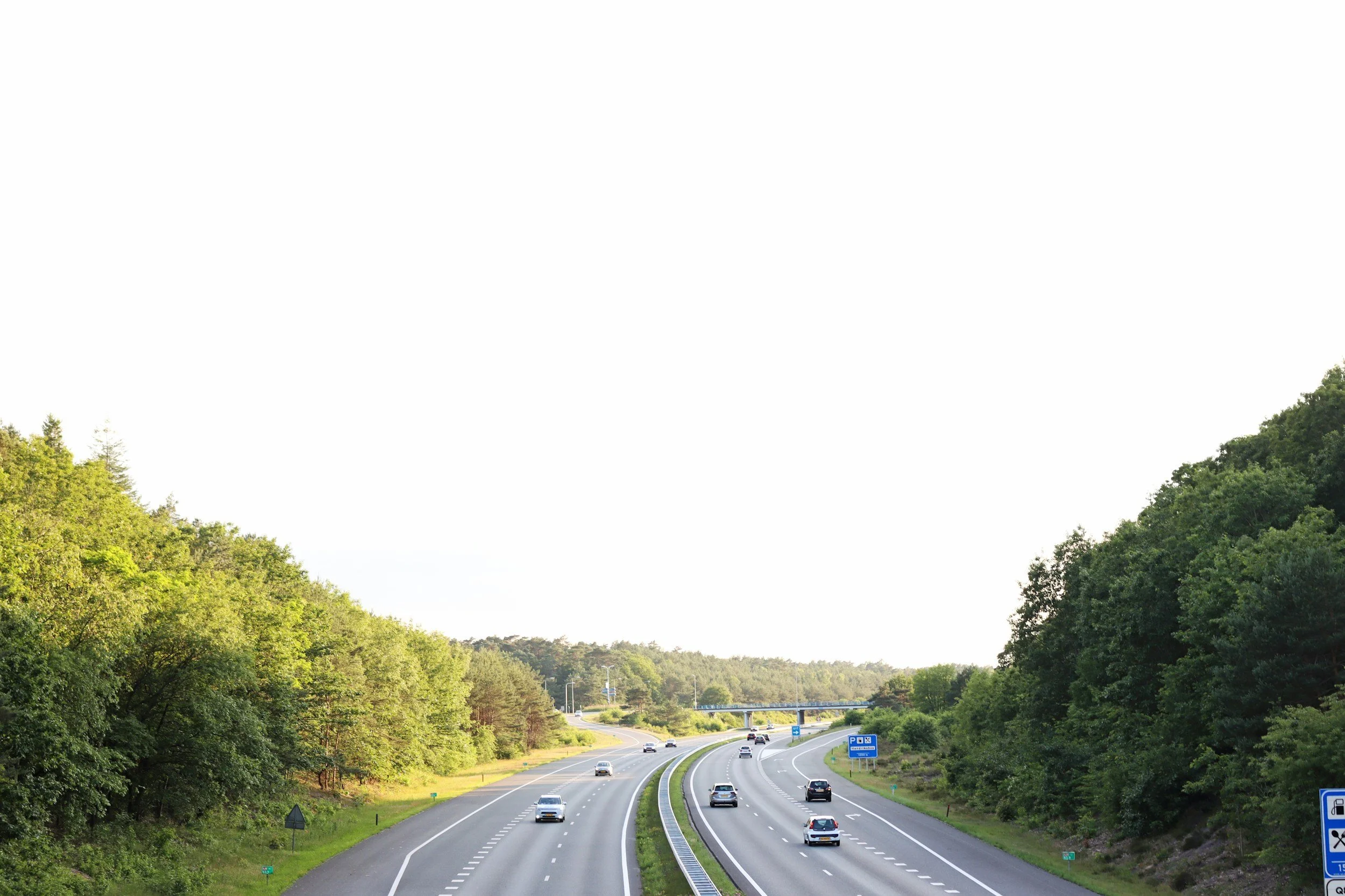 A highway with multiple lanes and cars driving in both directions, surrounded by trees and greenery on either side, under a clear sky.