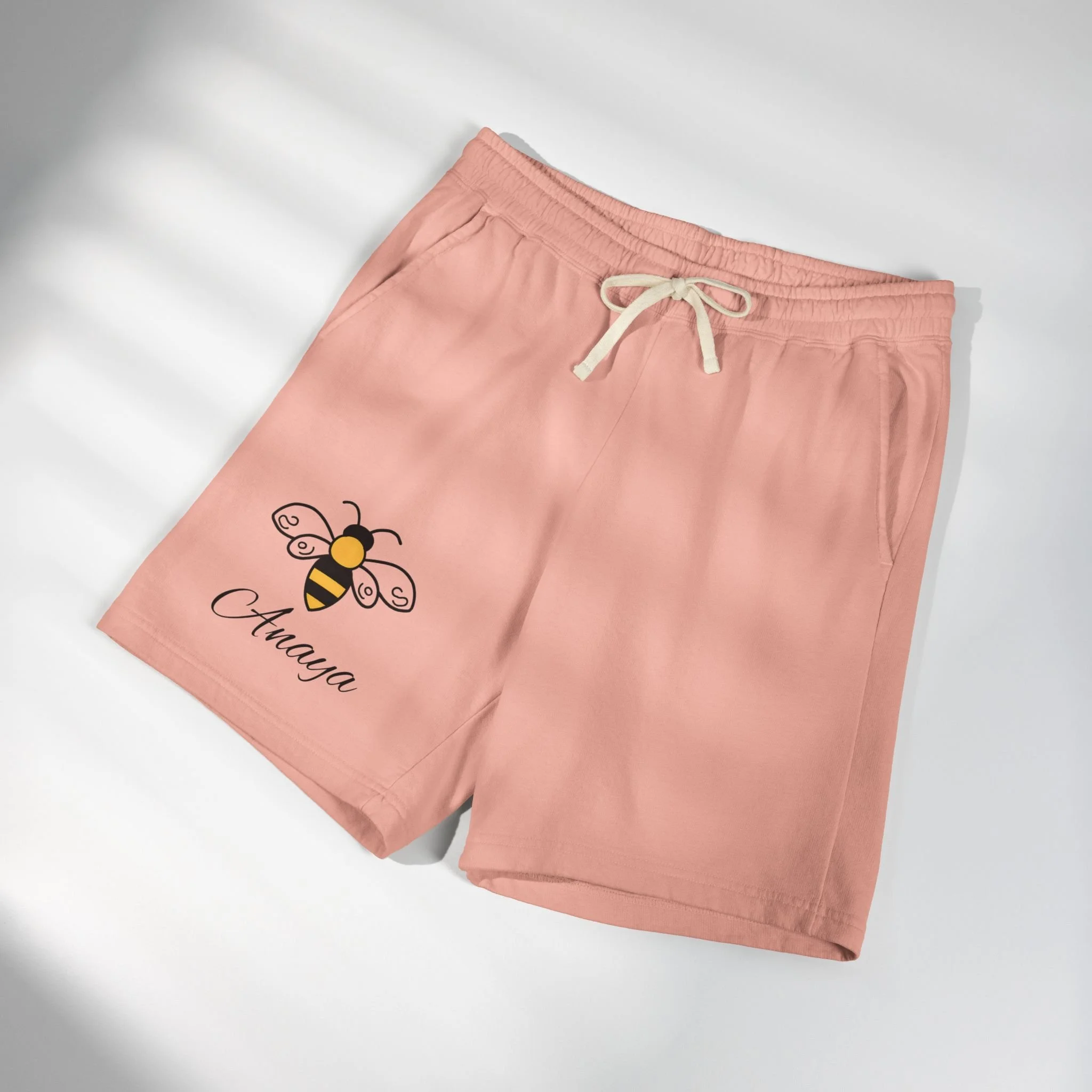 Pink drawstring shorts with a bee graphic and default name where personalization goes, laid out on a white surface.