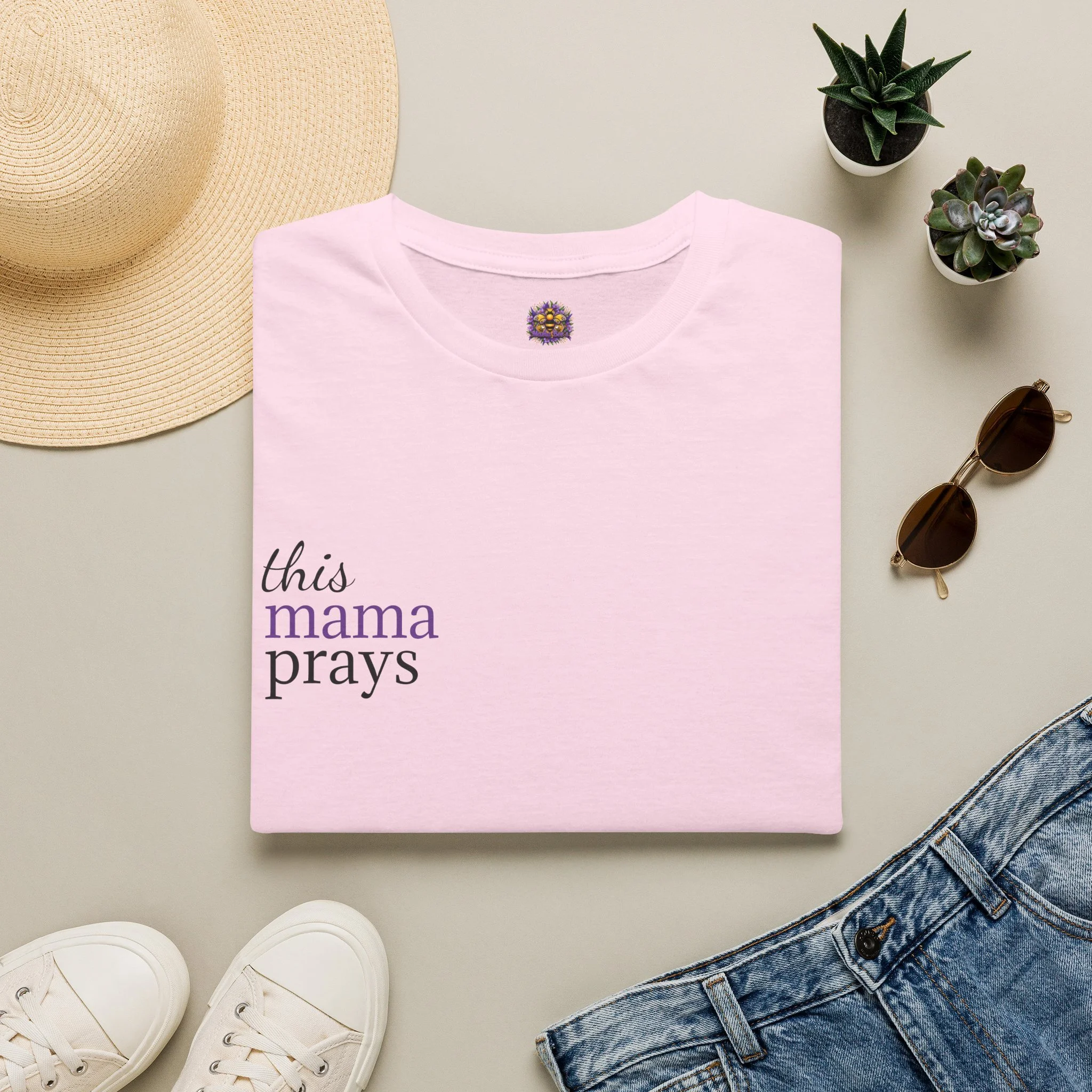 A flat lay of a beige woven sun hat, a pink T-shirt with Mama Bee's logo on the neckline, white sneakers, a pair of sunglasses, two small potted succulents, and denim jeans. Shirt reads "this mama prays".