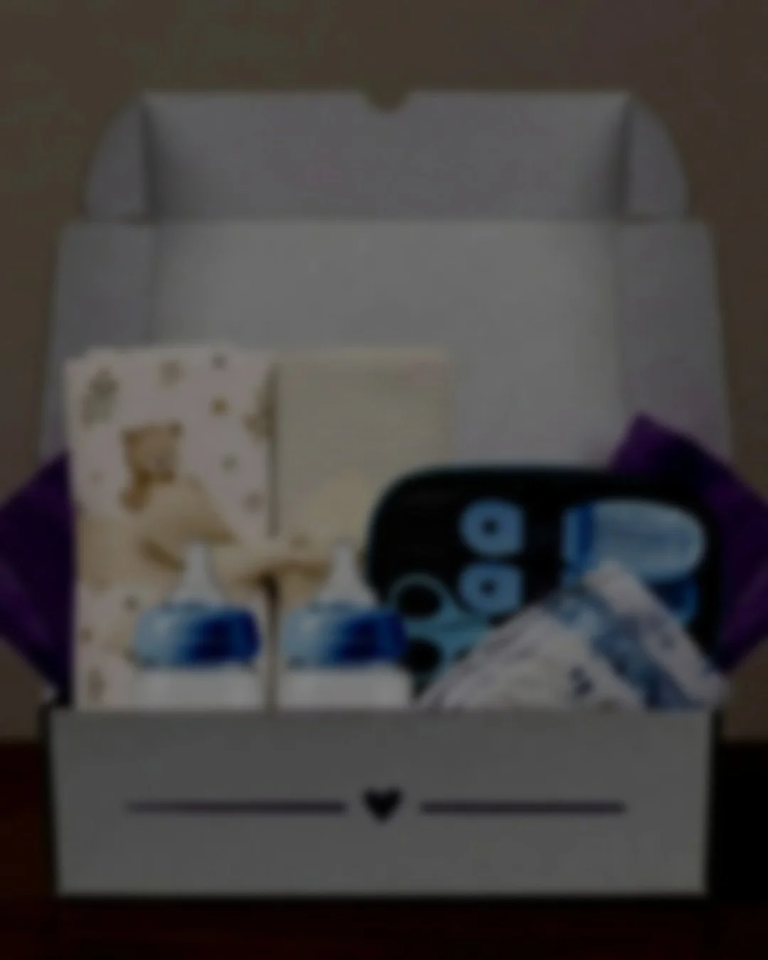 Open gift box containing baby items: two bottles, soft toy with bear design, striped items, baby scissors, brush, and napkins, decorated with purple tissue paper.