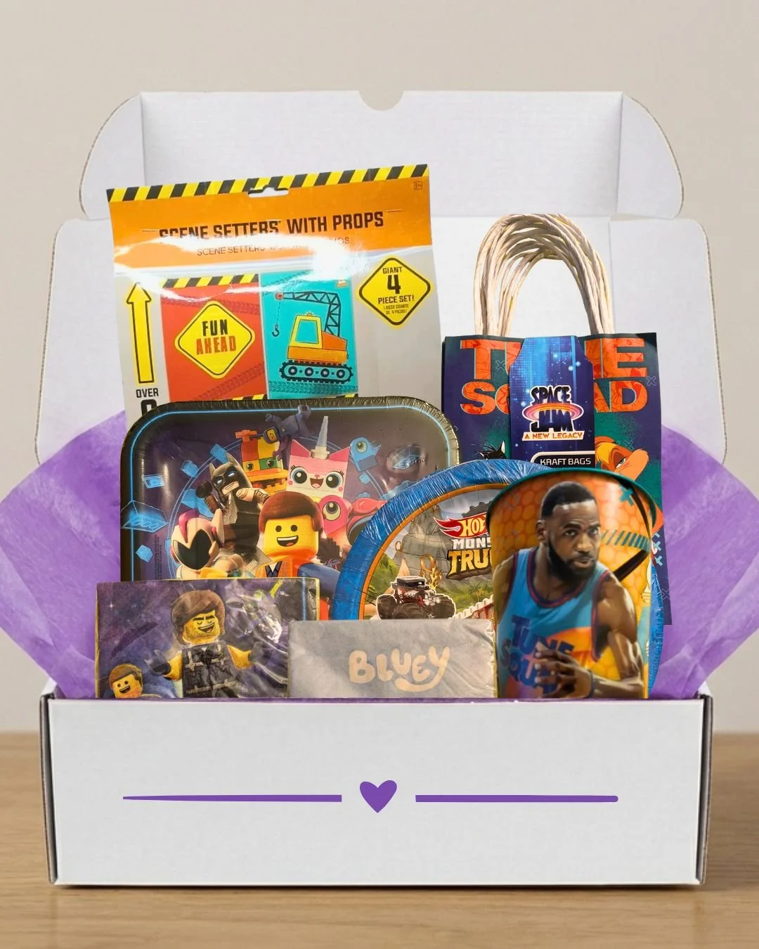 A gift box with purple tissue paper and a purple heart on the front, filled with various toys, snacks, and collectibles including LEGO figures, a Hot Wheels truck, a basketball player figurine, and themed snack packs.
