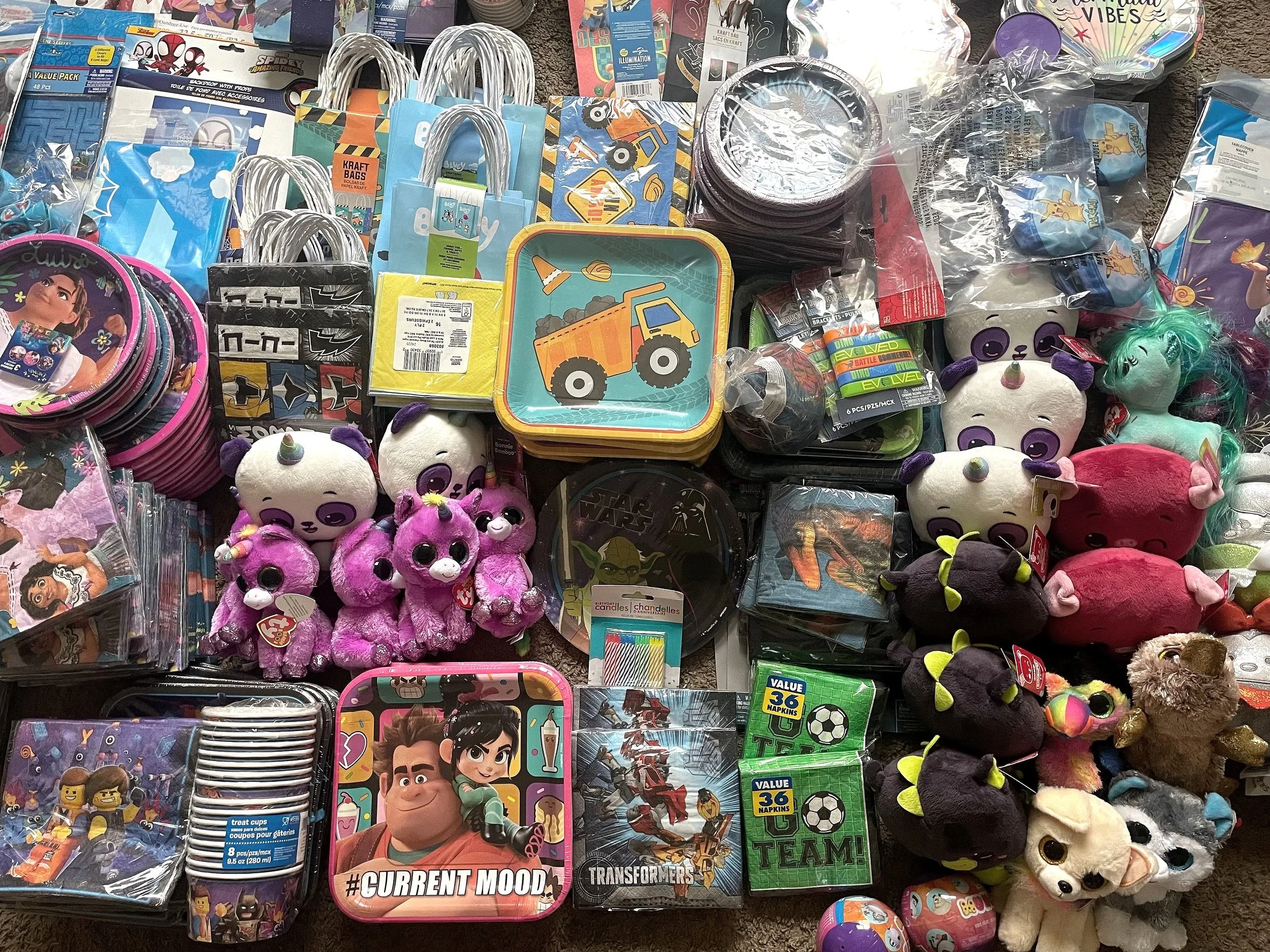 A wide variety of toys, plush animals, and gift bags spread out on a flat surface, featuring characters from popular cartoons and movies. Items such as plush pandas, unicorns, and superheroes are visible.