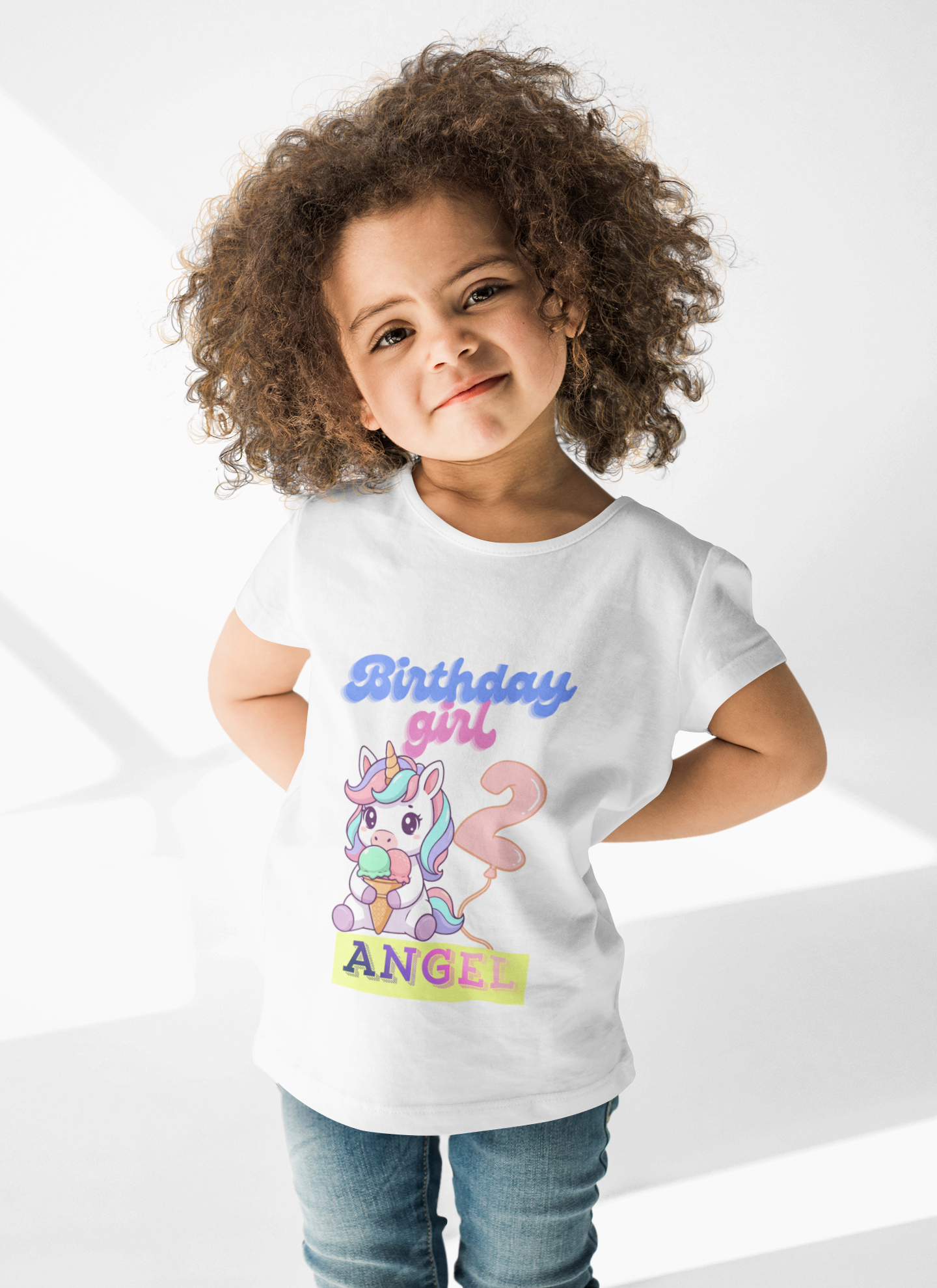 A young girl with curly hair smiling and posing, wearing a birthday girl t-shirt with a unicorn and the words 'Birthday girl' and 'Angel'. She is standing against a white background.