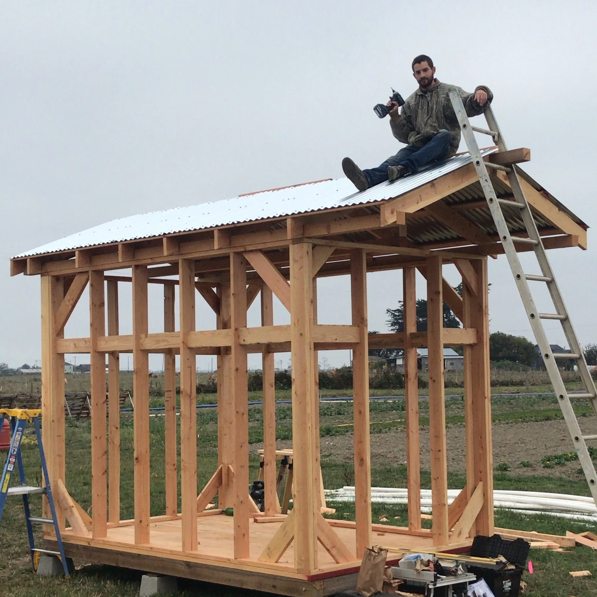 A man sitting on the roof of a small wooden structure under construction, holding a drill, with a ladder leaning against the building.