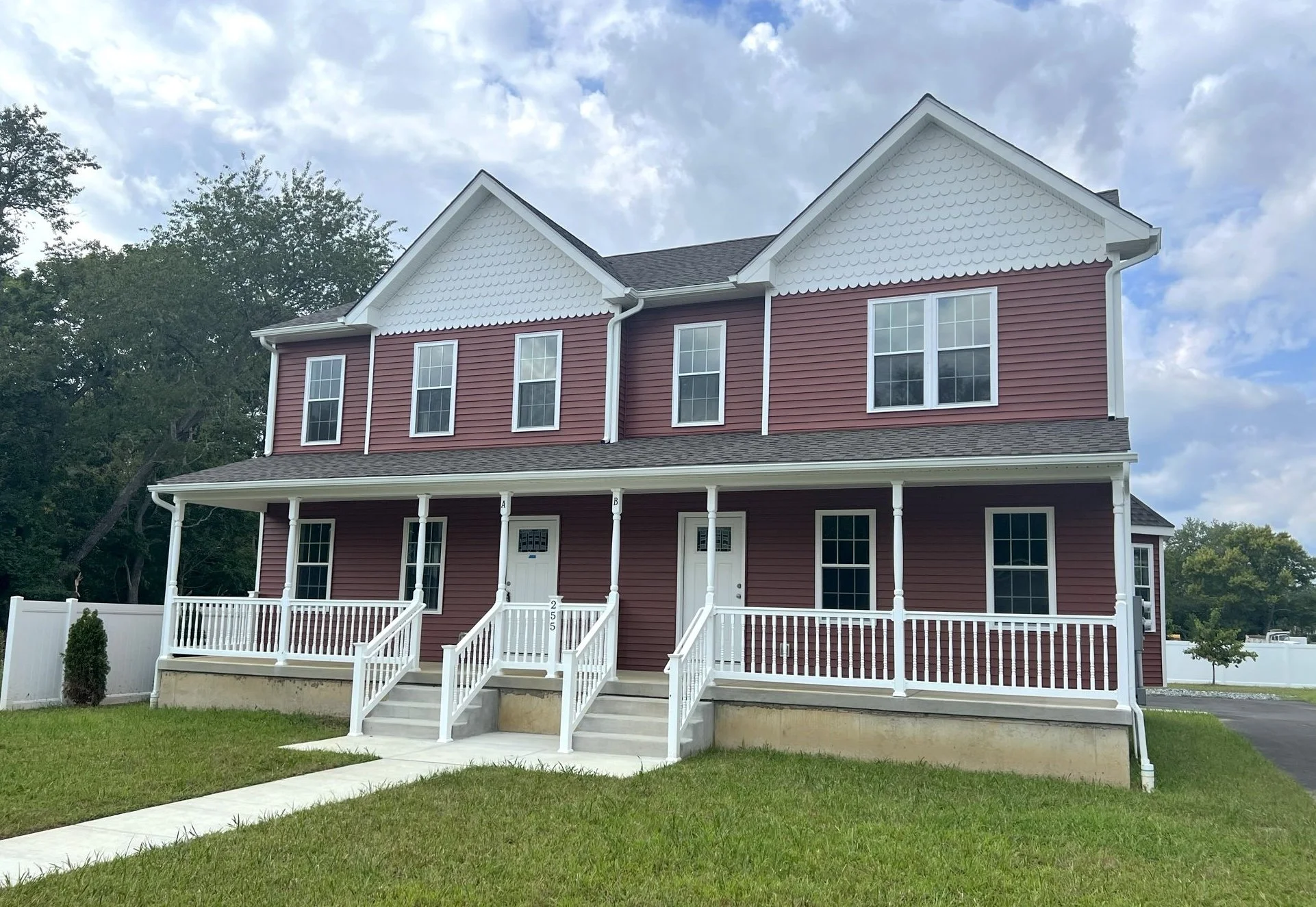 A two-story red house with white trim, large front porch with white railings, and several tall windows, surrounded by green grass and trees under a partly cloudy sky.