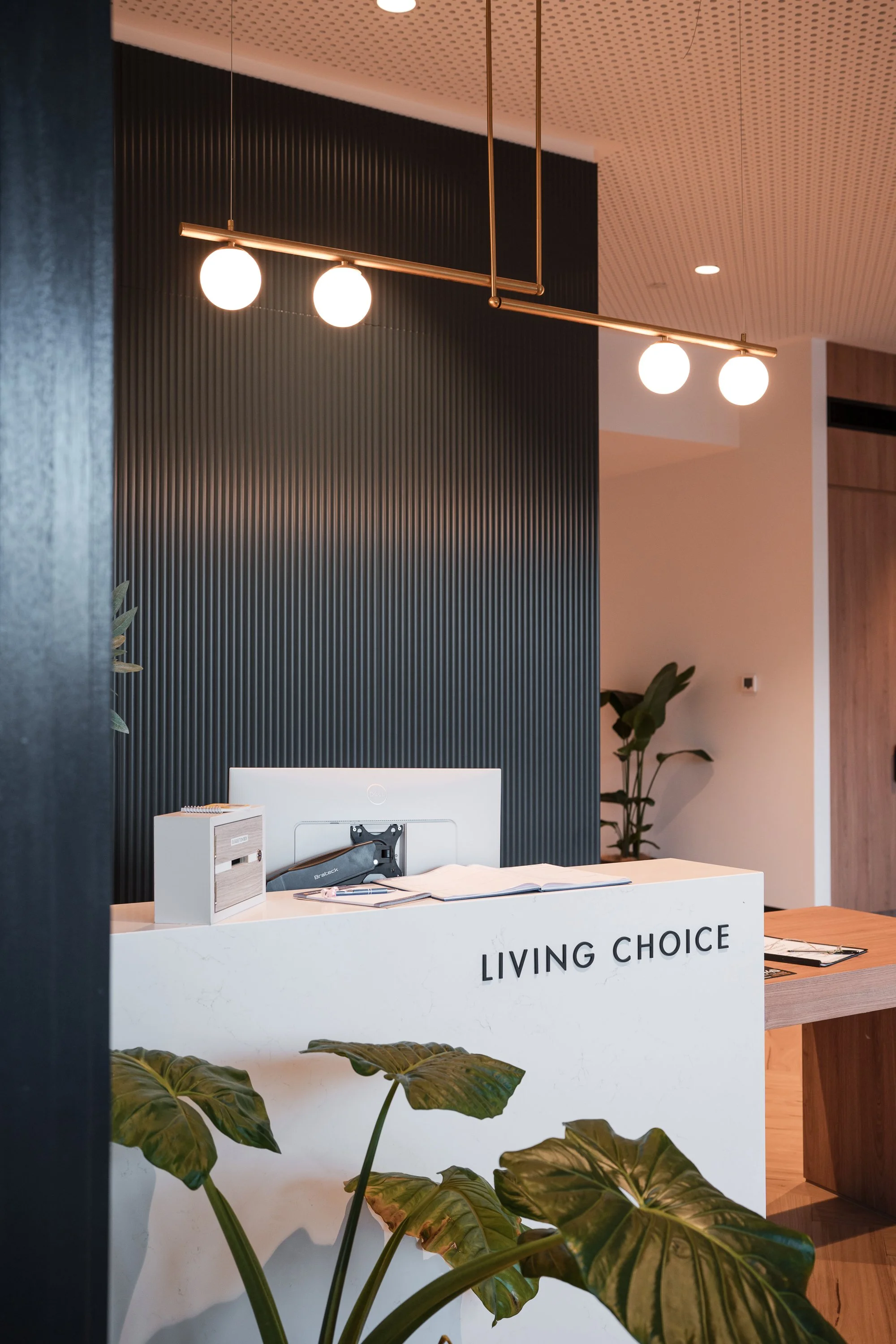 Modern reception desk with the words 'Living Choice' on it, surrounded by green plants, with a black textured wall and wood accents in the background. Overhead lighting features a minimalist gold fixture with three globe lights.