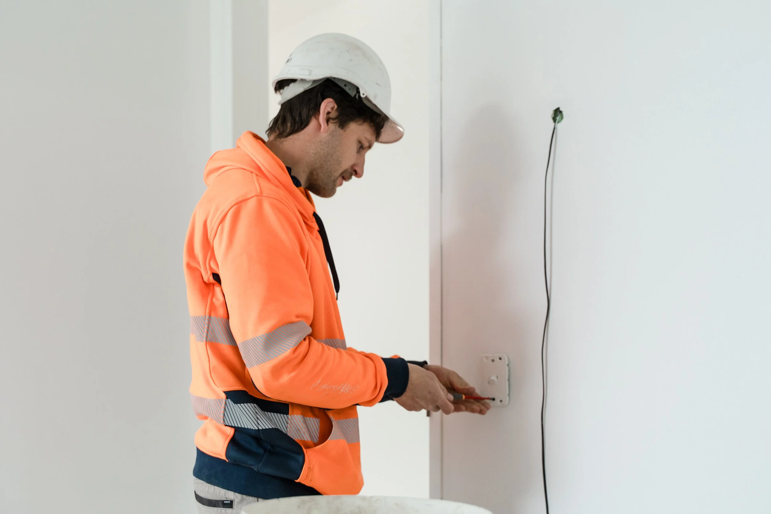 A construction worker in an orange safety jacket and white helmet is installing an electrical outlet on a white wall.