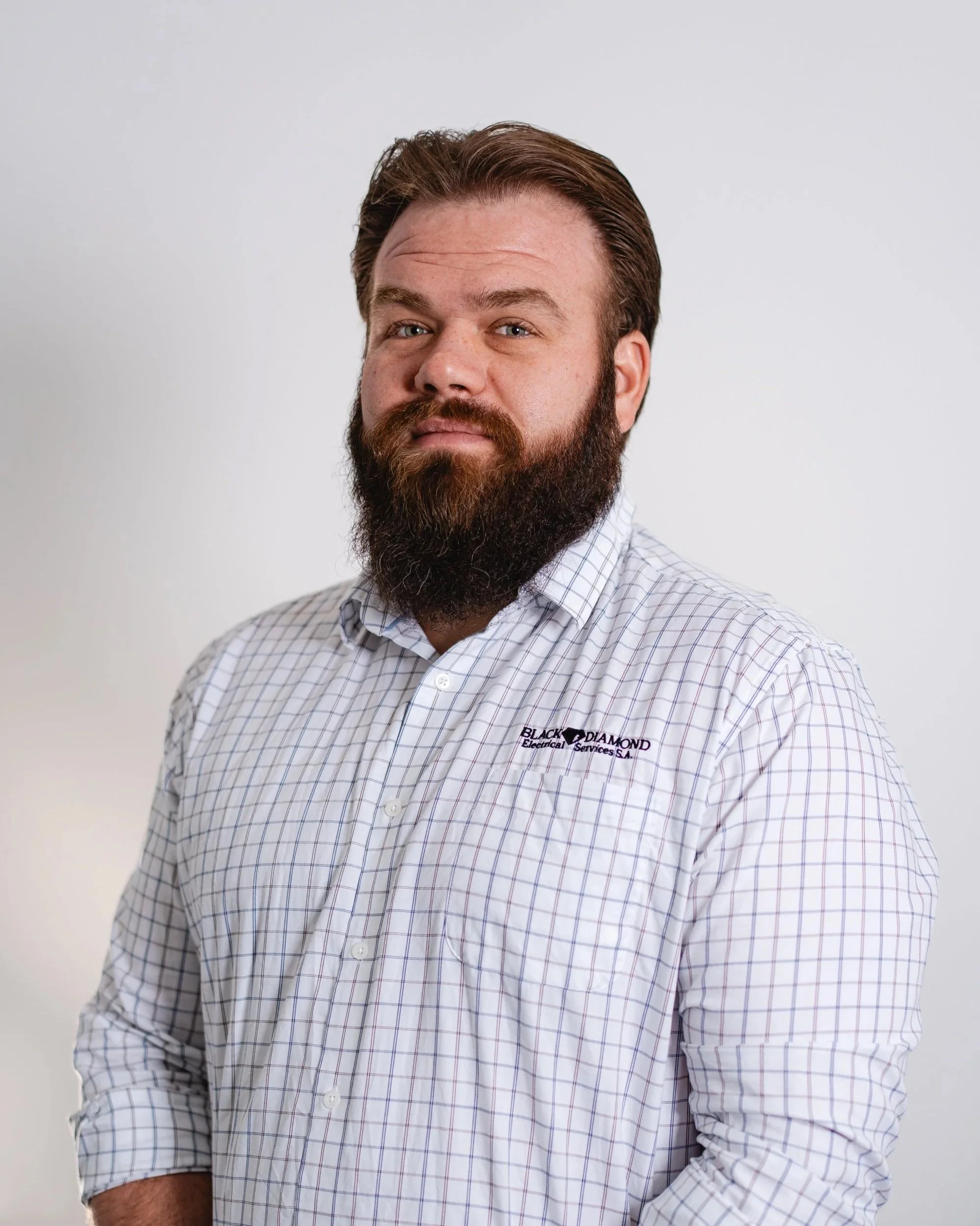 Portrait of a man with a beard and mustache wearing a checkered shirt with company logo, standing against a plain white background.