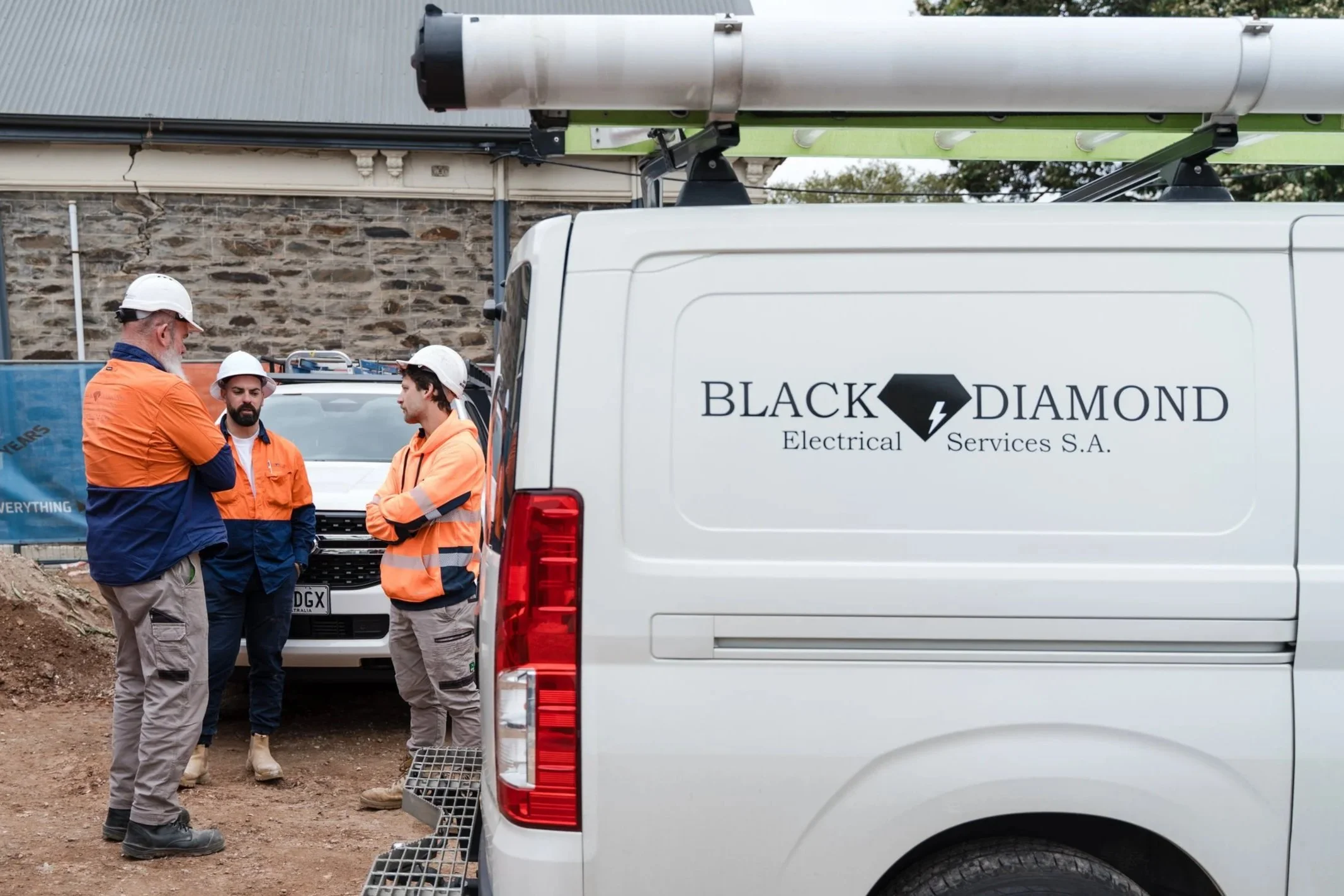 Three electricians wearing safety helmets and orange jackets having a discussion next to a white van with company branding, parked near a construction site with a brick wall in the background. The van is equipped with a ladder on the rear.
