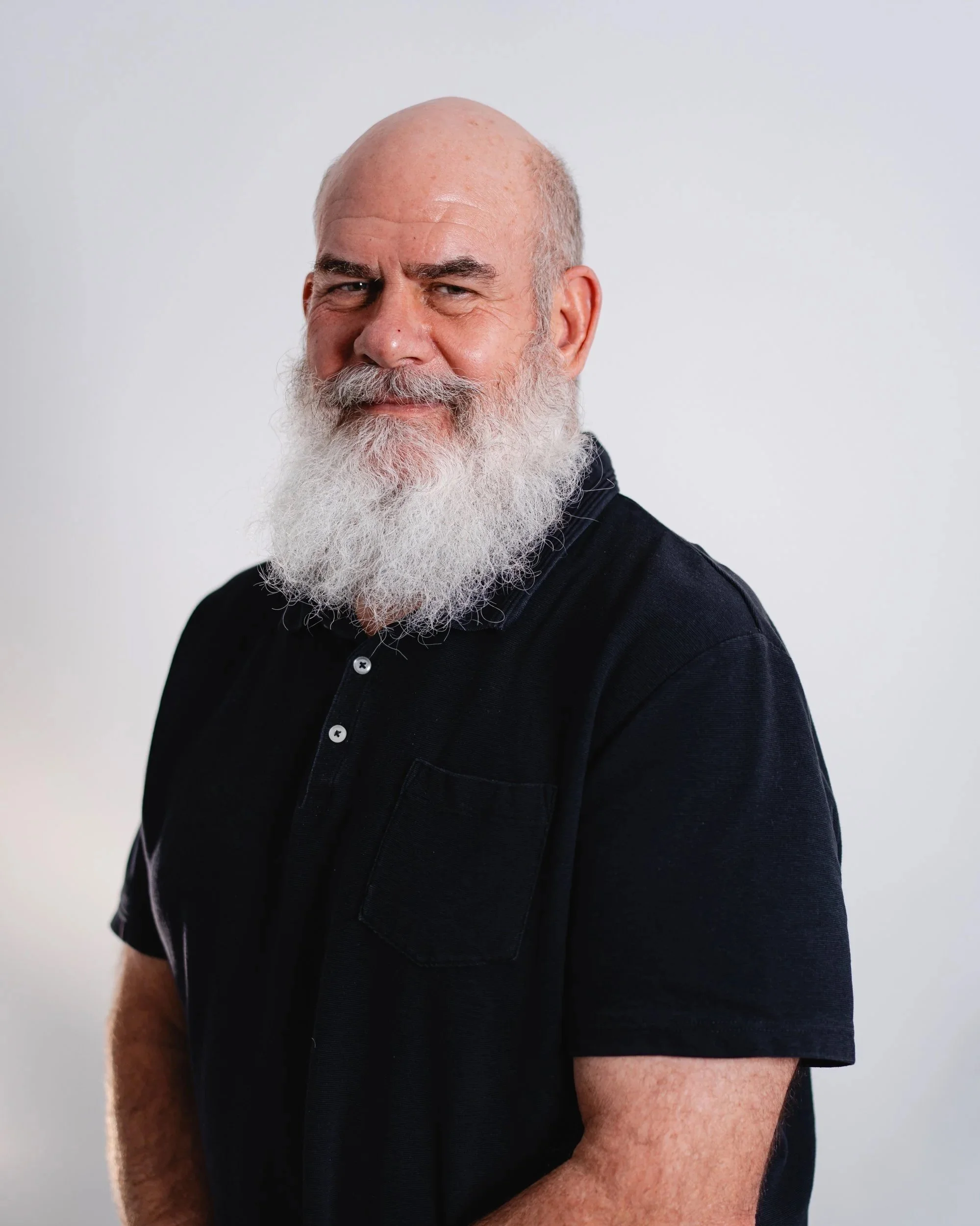 Portrait of a bearded man with a bald head, wearing a black shirt, standing against a plain white background.