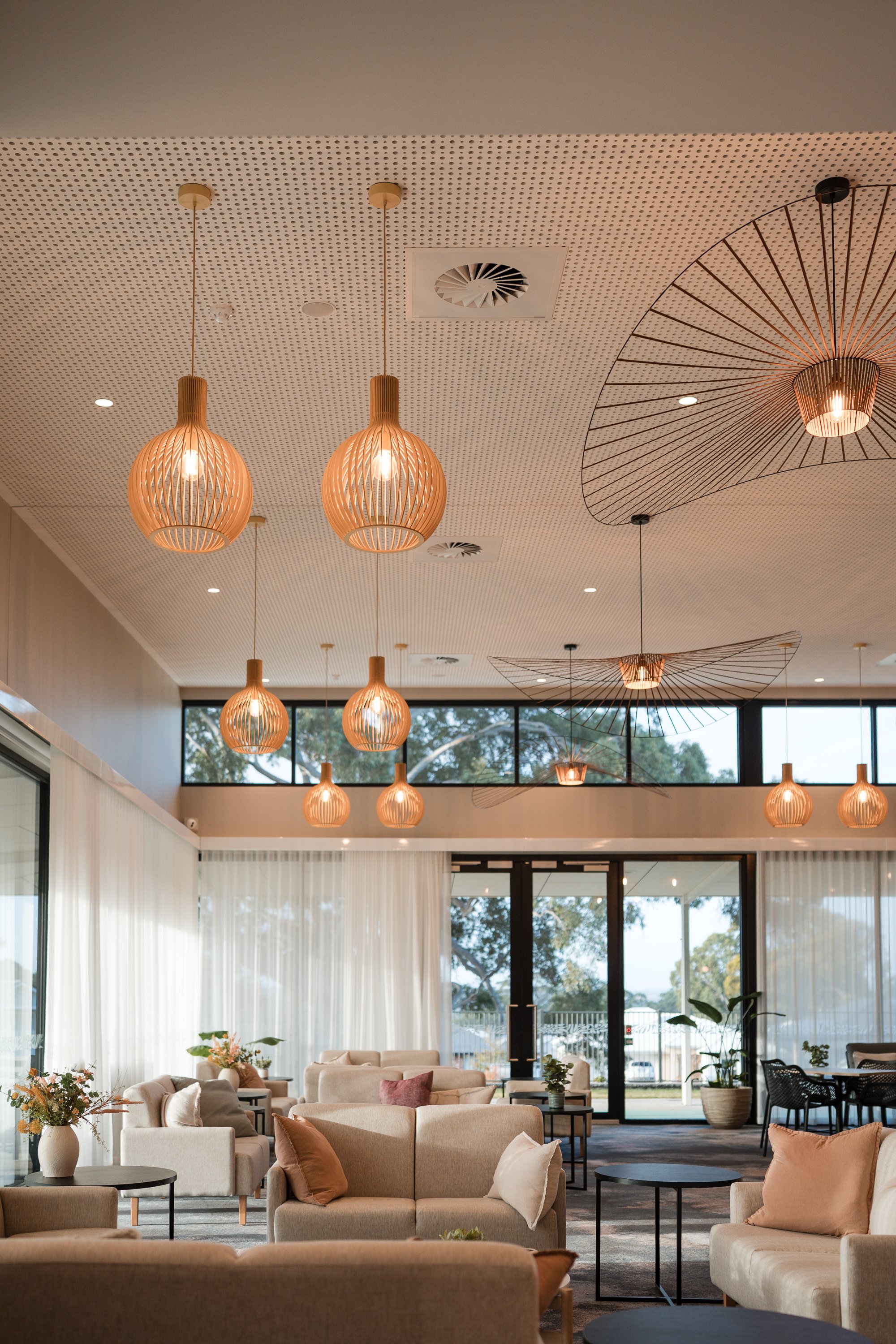 Modern lounge with beige sofas, side tables, and large windows, decorated with potted plants and flowers, with hanging wooden and wire-frame pendant lights from the ceiling.