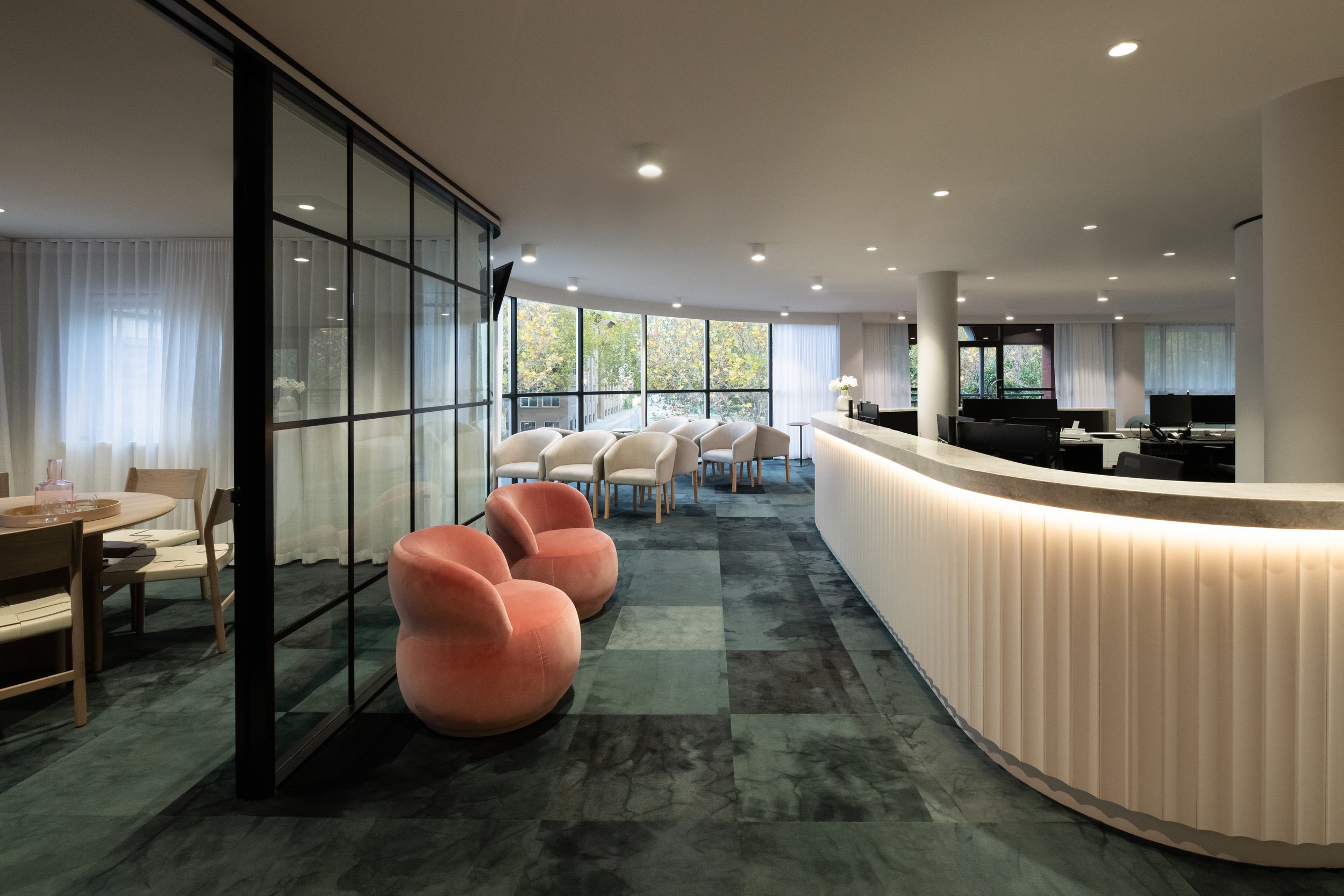 Modern reception area with curved white desk, plush pink chairs, beige seats, and large windows with white curtains and greenery outside.