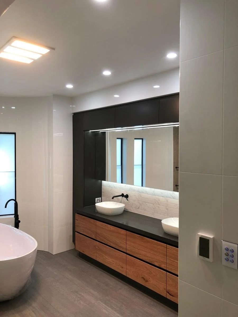 Modern bathroom with a large mirror, two vessel sinks, wooden cabinet drawers, bathtub, and a white tiled wall