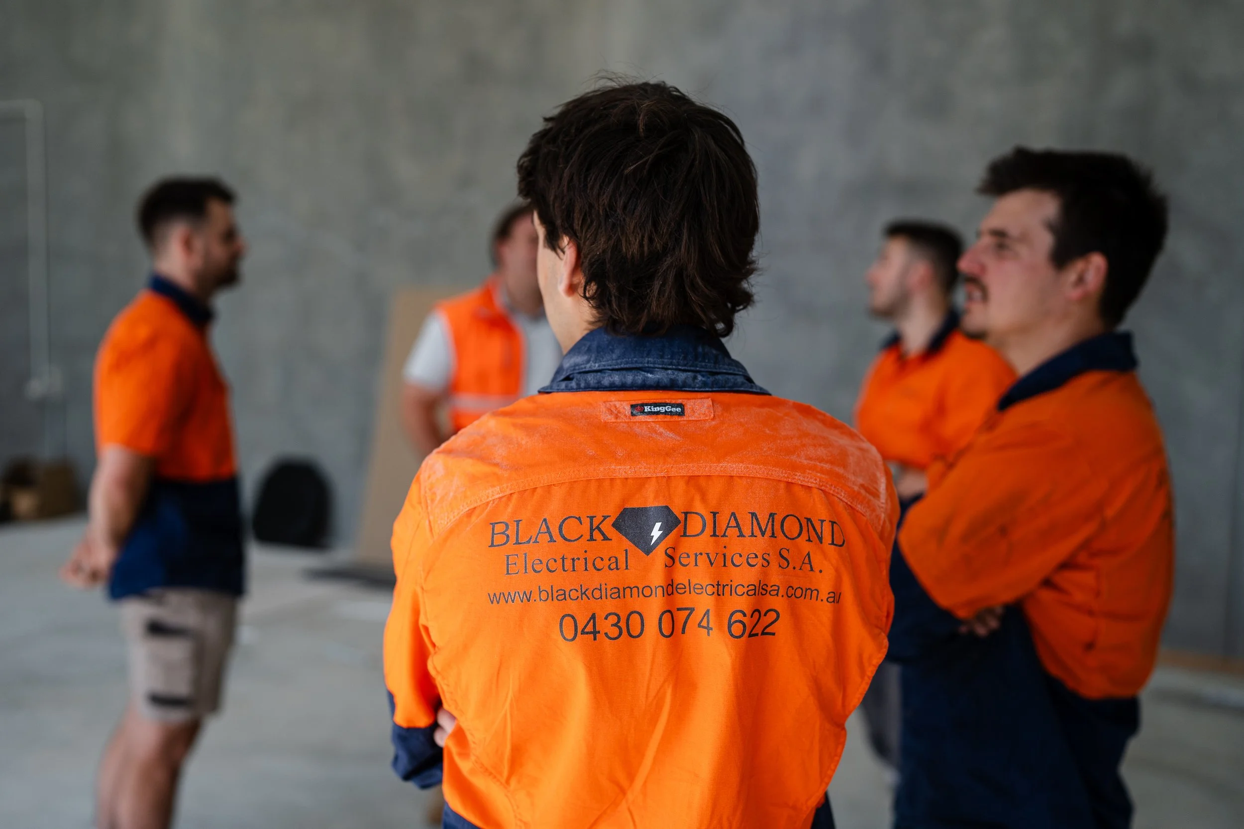 A person with short brown hair wearing an orange safety vest with black text and a logo, standing in a group of four people inside a room with plain gray walls, engaging in a discussion.
