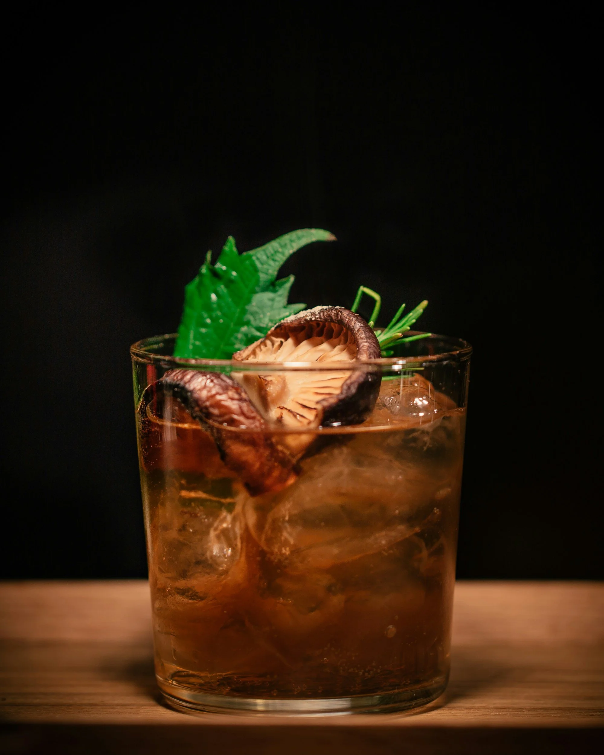 A glass of whiskey with ice, garnished with a mushroom, a green leaf, and a sprig of rosemary, on a wooden surface against a black background.