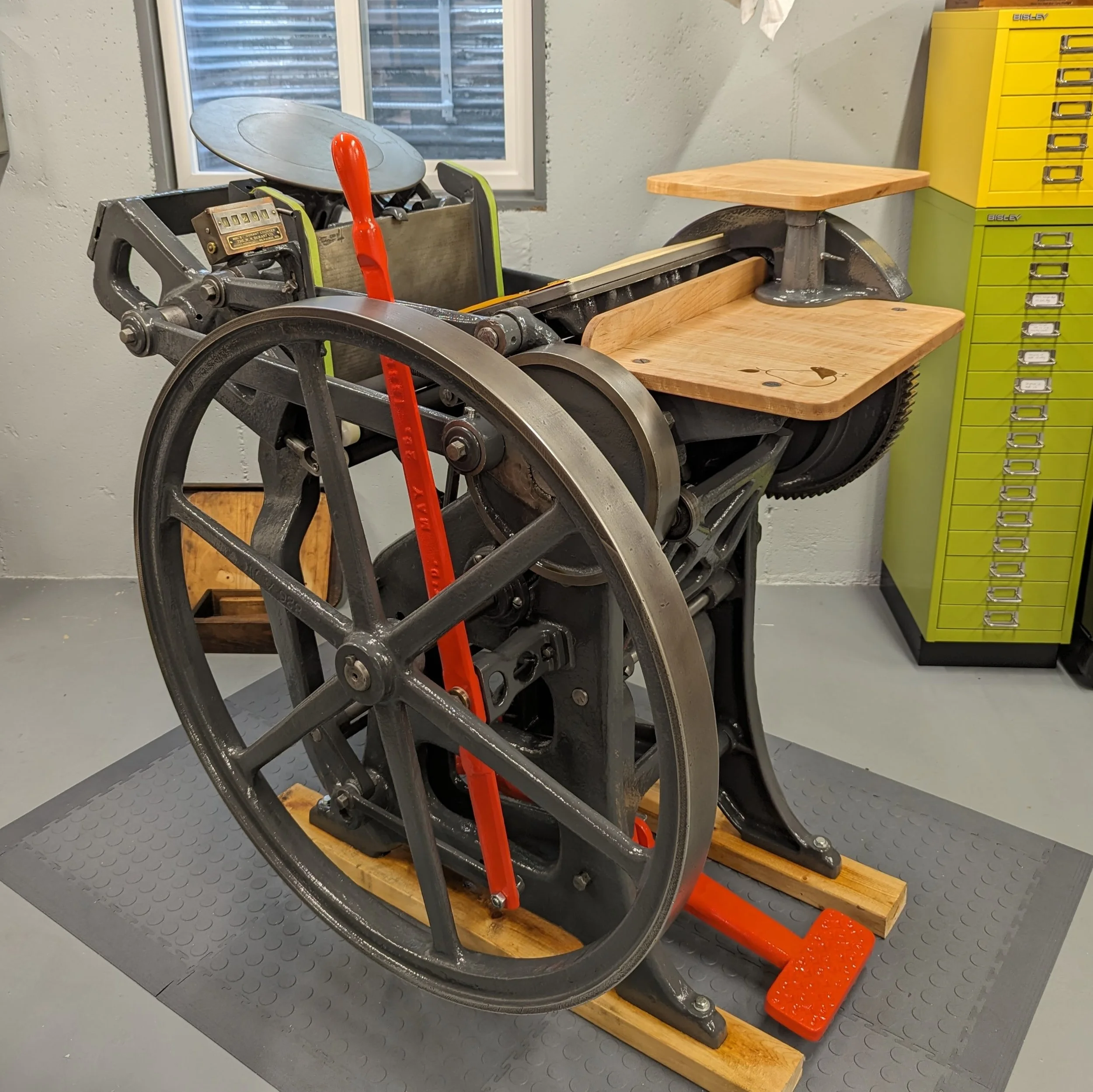 Vintage hand-fed, treadle operated, letterpress printing press with large fly wheel and wood feed and delivery tables , in a workshop, with window and yellow storage cabinet in the background.