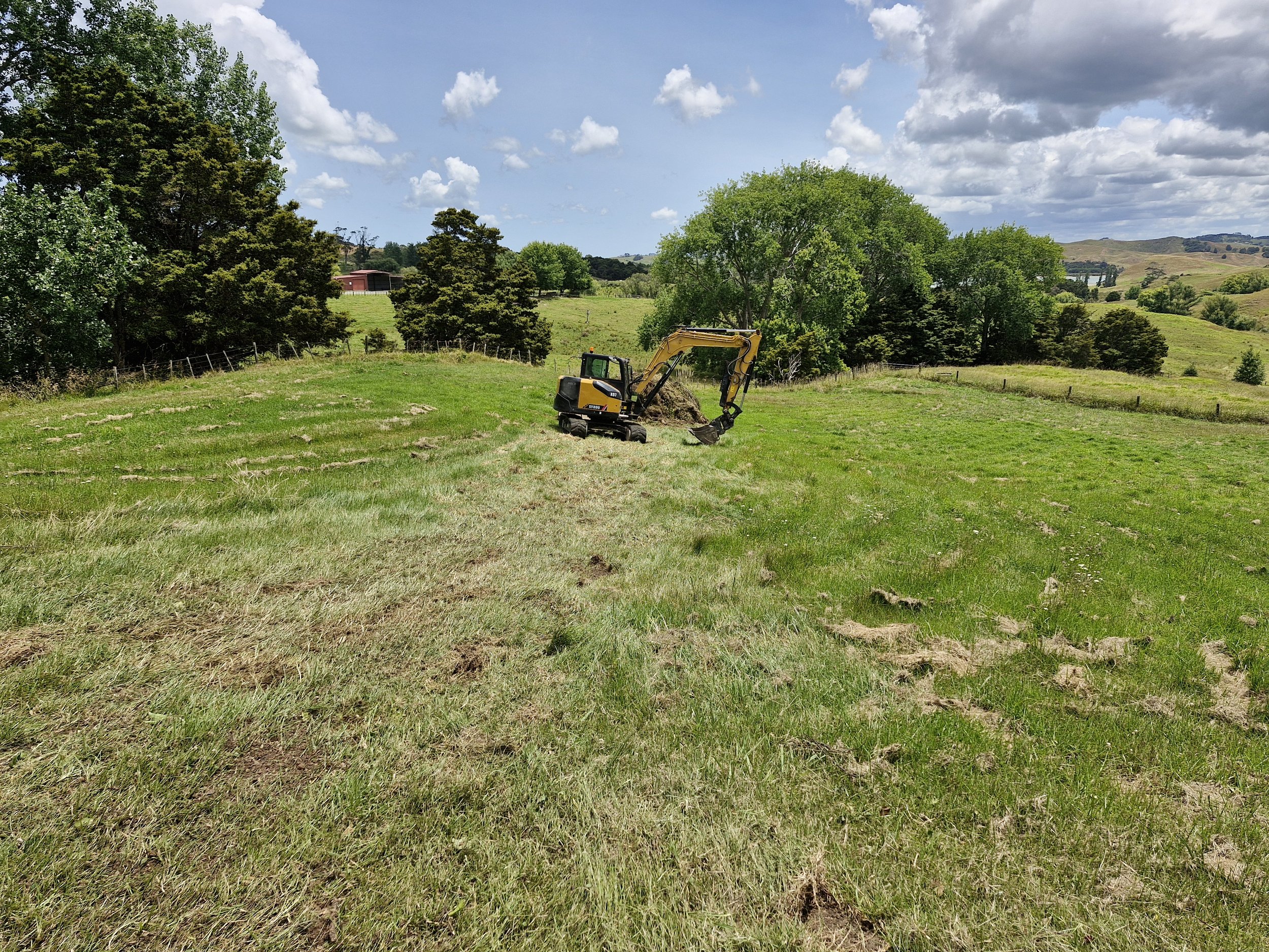 A small yellow and black excavator working on a grassy field under a partly cloudy sky with trees and hills in the background.