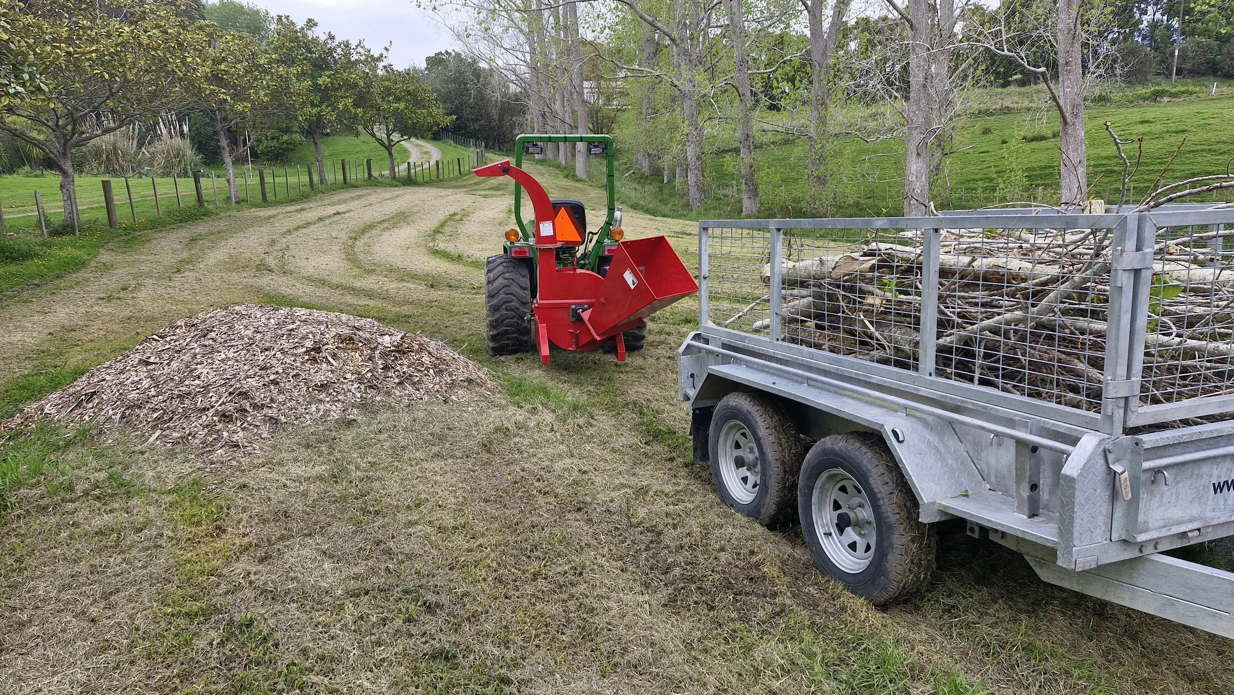 A small red wood chipper attached to a green tractor, with a trailer filled with branches and wood chips on a grassy farm path surrounded by trees and fencing.