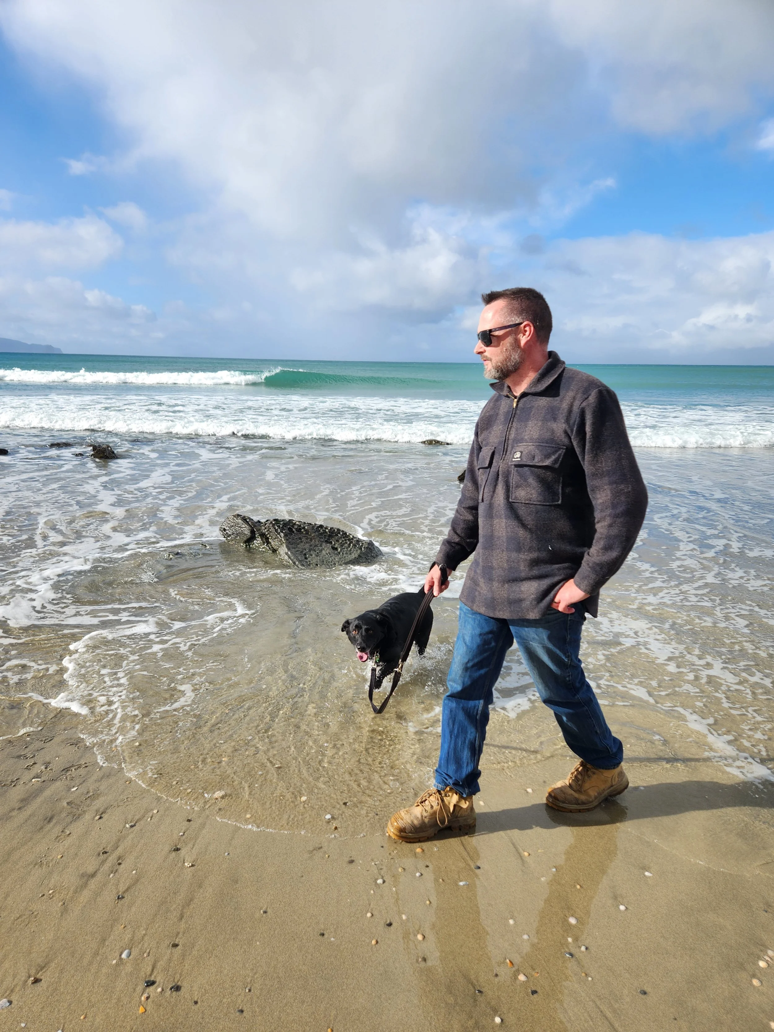 James Milne walking his black dog, Gus, along Mangawhai Beach beach with sand, rocks, and ocean waves, under a partly cloudy sky.