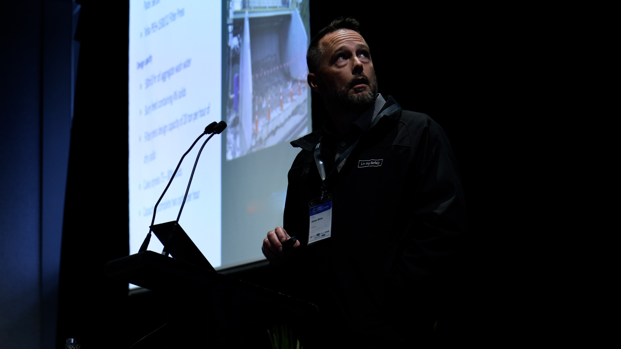 James stands at a podium with a microphone, looking up at a large projection screen displaying presentation slides with text and images.