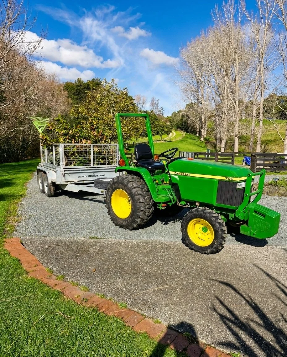 Green tractor pulling a trailer filled with plants on a gravel driveway in a rural area with trees and a blue sky.