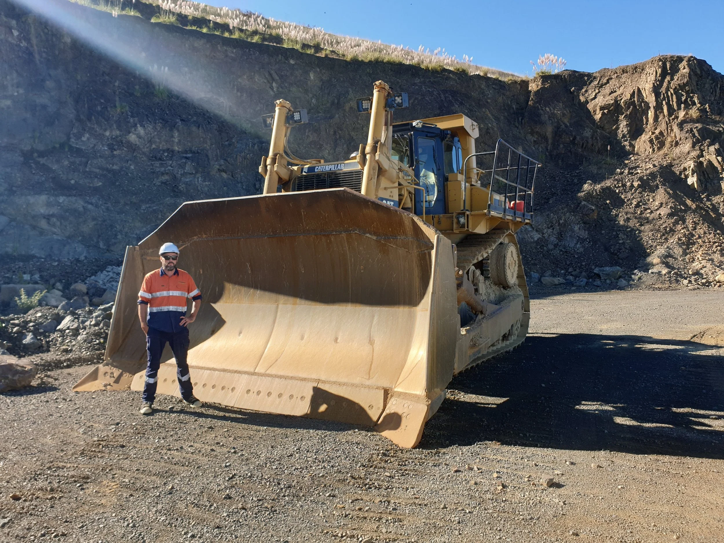 James wearing safety gear, including a white hard hat and an orange and blue safety uniform, standing in front of a large yellow bulldozer on a gravel surface at a construction site surrounded by rocky terrain.
