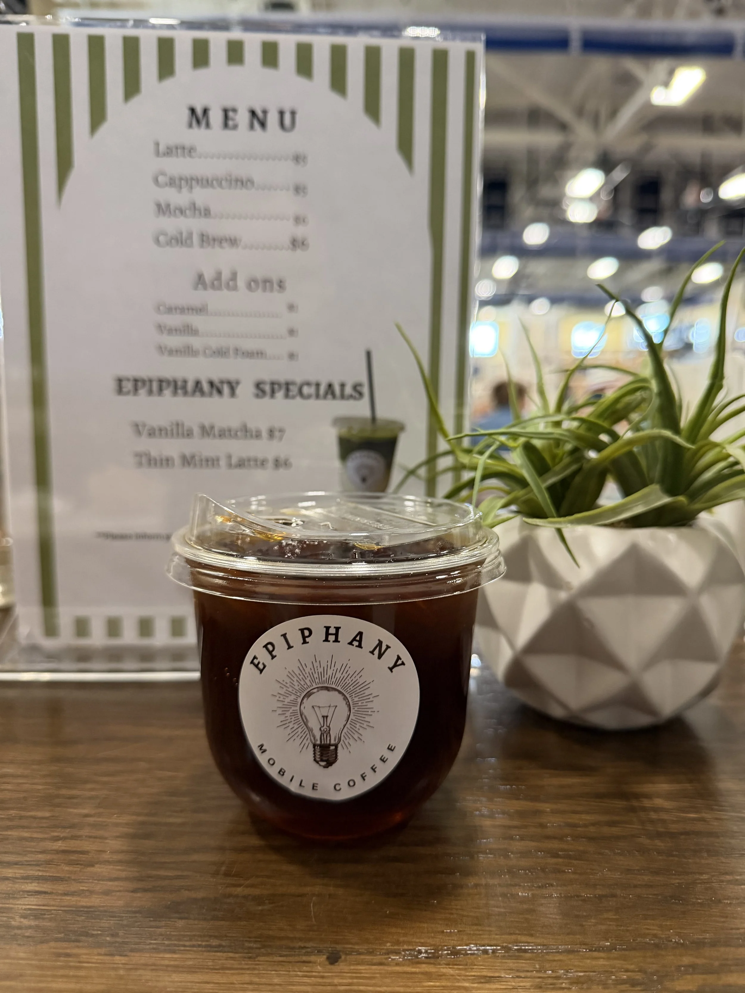 Iced black coffee in a plastic cup with Epiphany logo, behind a menu and a potted plant on a wooden table at a coffee shop.