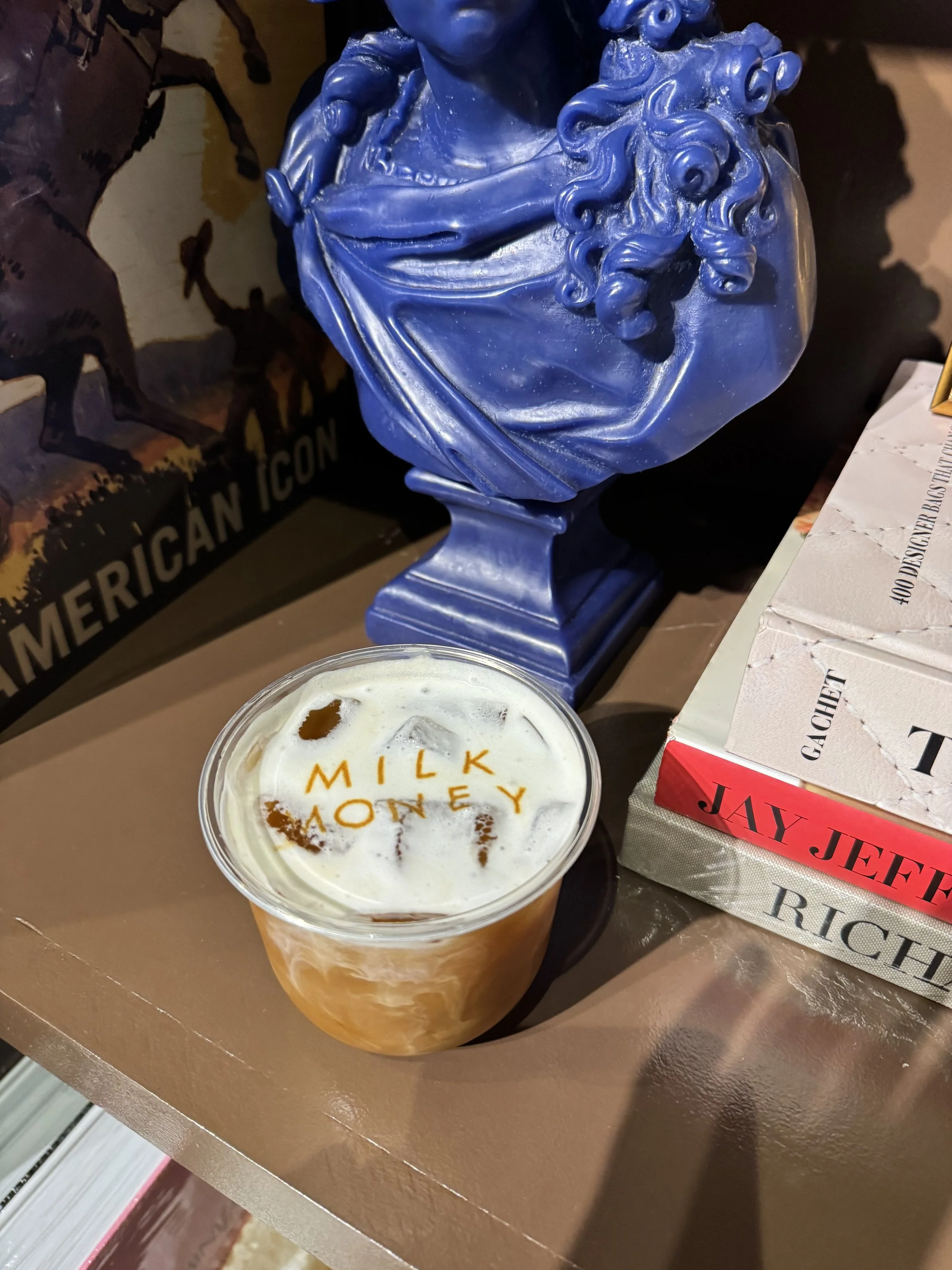 A blue sculpture bust of a woman with curly hair and draped clothing, placed on a table. In front, a cup of drink with the words "milk" and "money" written on the foam. Part of a boxed item, an American Icon poster, and a book are also visible on the