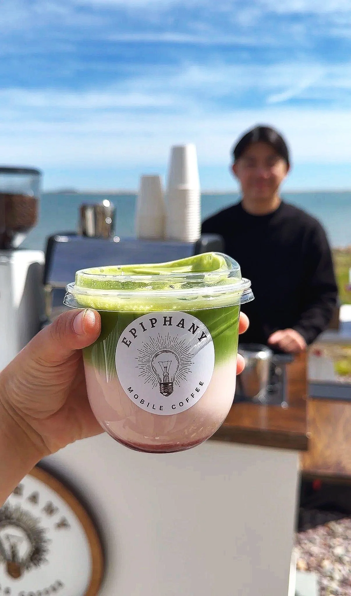 A hand holding a cup of pink and green layered coffee from Epiphany Mobile Coffee, with a blurred background of a person and outdoor setting including a clear blue sky and ocean.