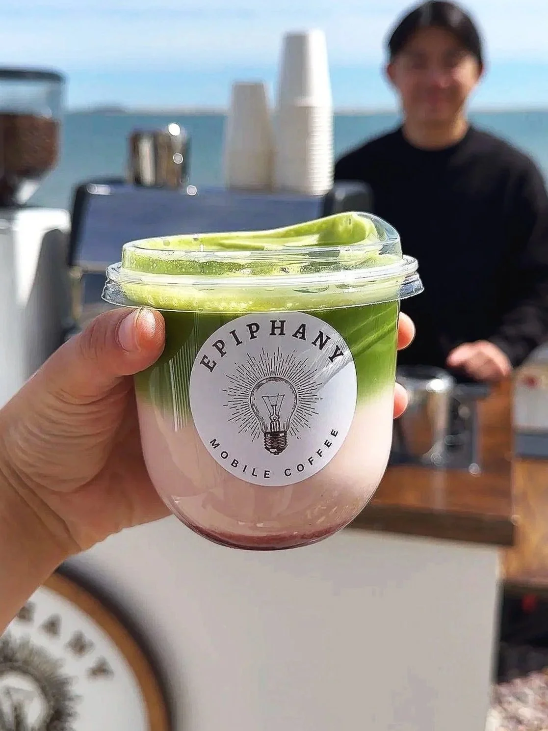 Hand holding a pink and green layered canned beverage with a logo that says 'E Piphany Mobile Coffee' in front of a person wearing black at an outdoor coffee stand by the ocean.