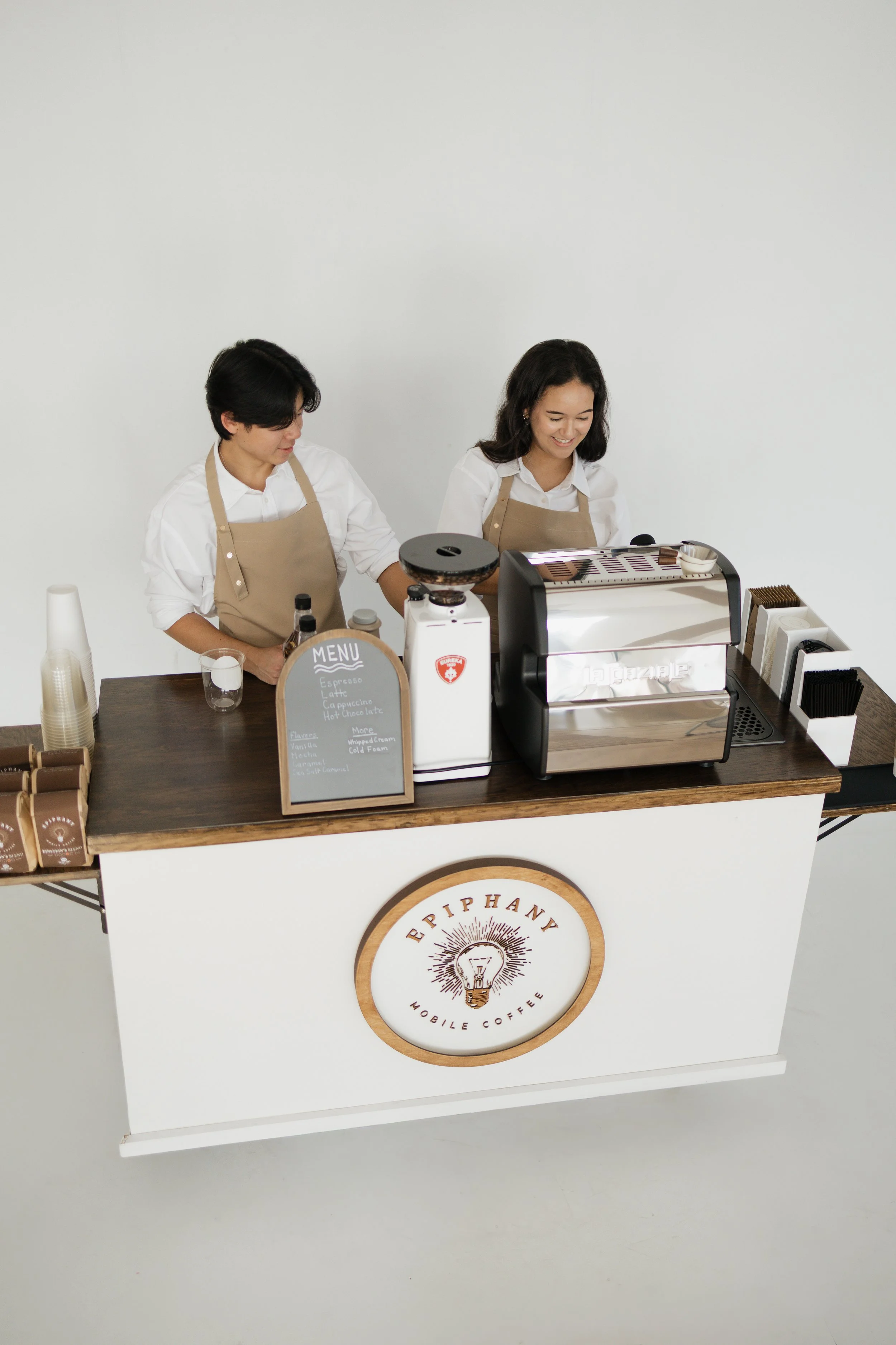 Two baristas standing behind a mobile coffee shop counter with a sign that reads "Epiphan Mobile Coffee," and various coffee-making equipment.
