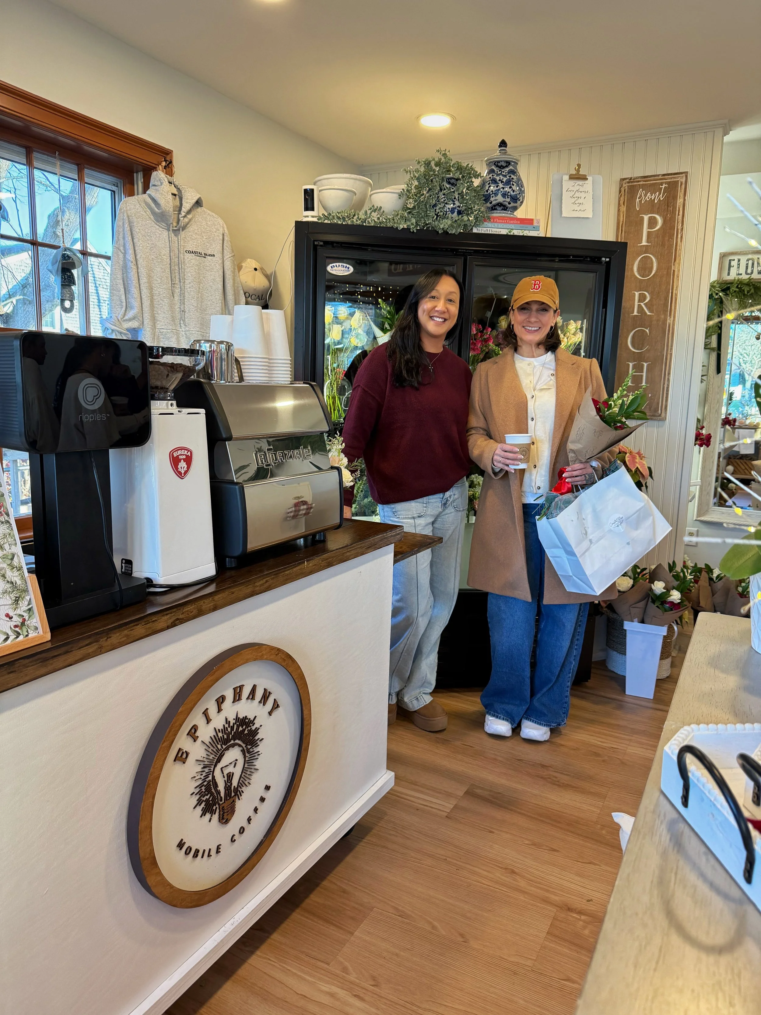 Two women standing inside a coffee shop near the counter, one holding a bouquet of flowers and a gift bag, both smiling, with coffee equipment and decorative items in the background.
