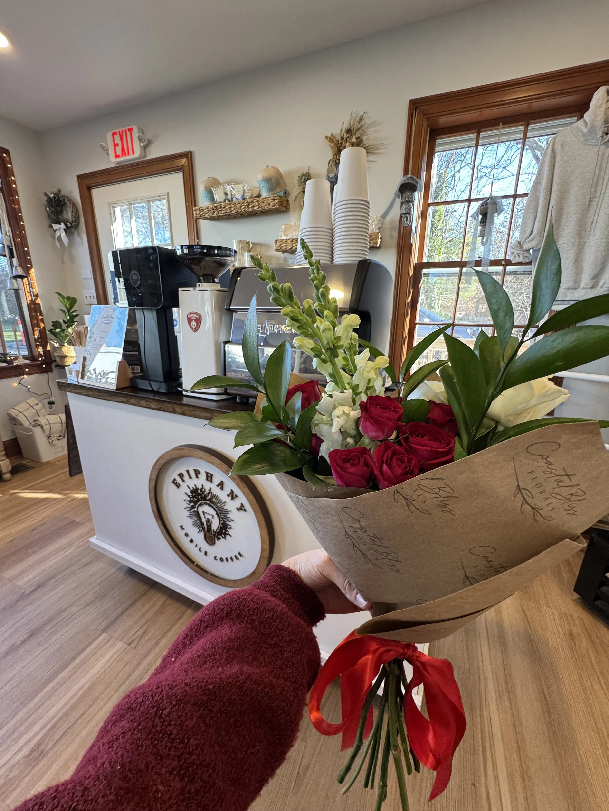 Person holding a bouquet of red and white flowers wrapped in paper inside a coffee shop with a white counter and coffee machine, and a window showing trees outside.