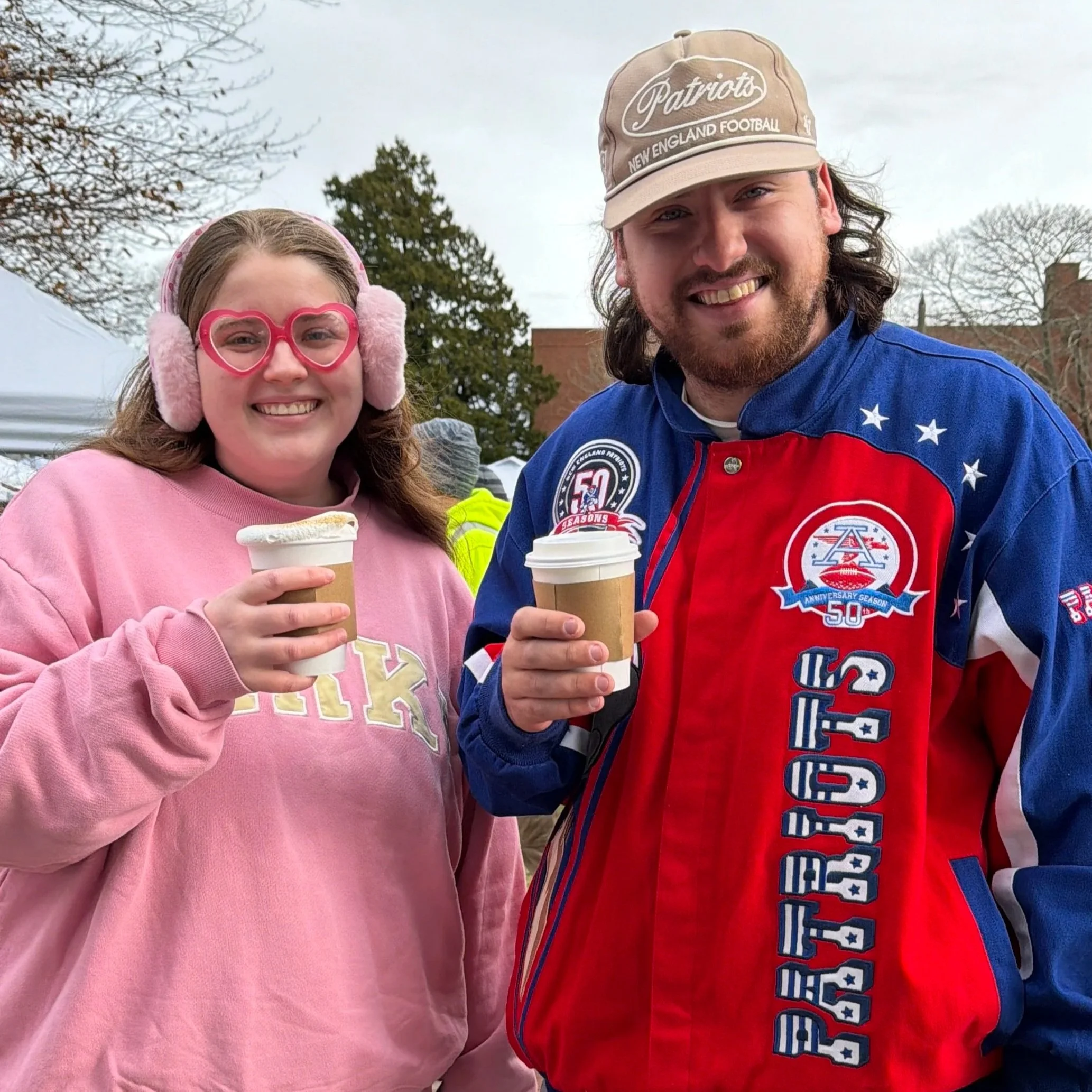 A woman and a man smiling and holding cups of hot chocolate outdoors. The woman is wearing pink glasses shaped like hearts, pink earmuffs, and a pink sweatshirt. The man is wearing a beige Patriots cap and a red, white, and blue jacket with patriotic