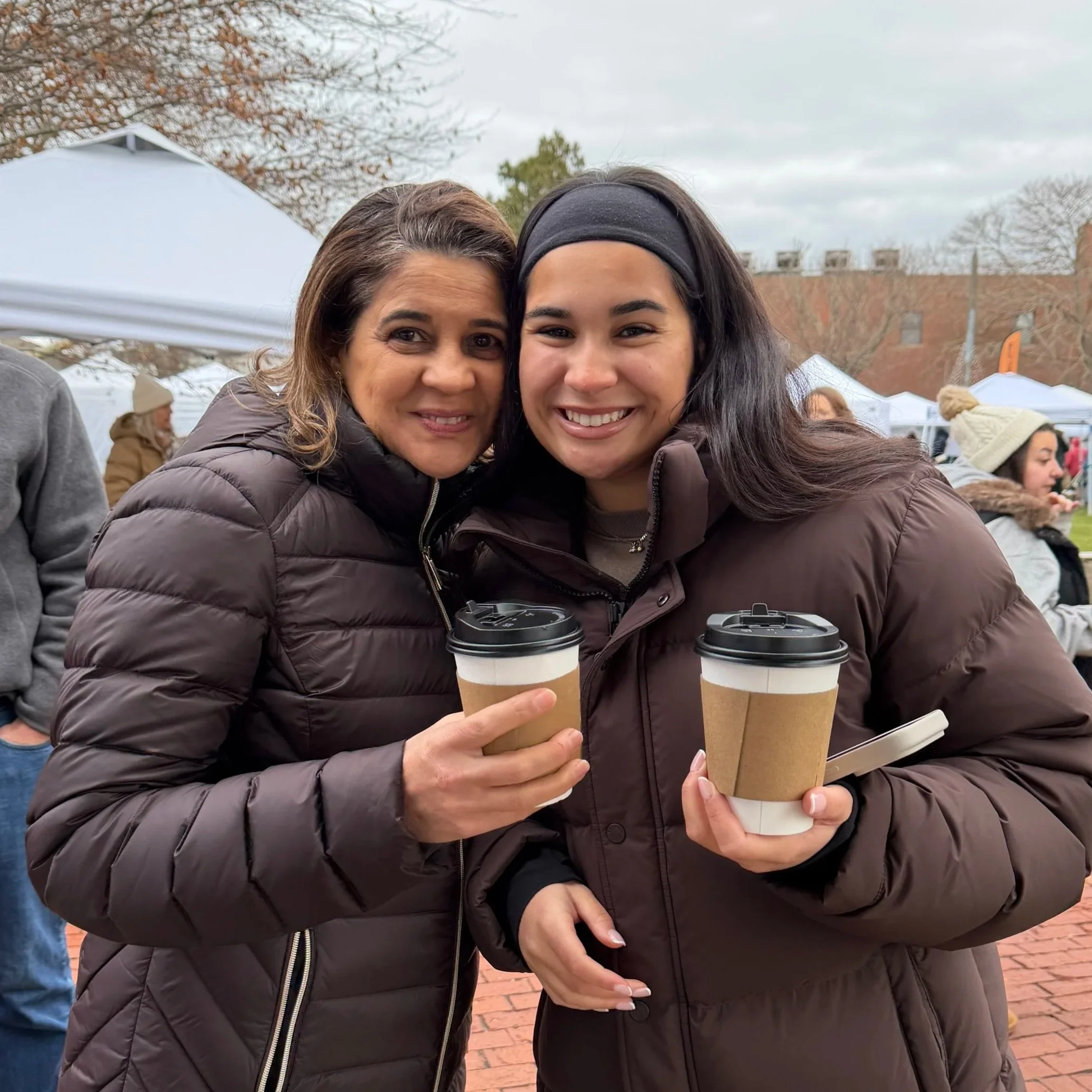 Two women smiling at an outdoor event, holding coffee cups, wearing brown puffer jackets, with tents and people in the background.