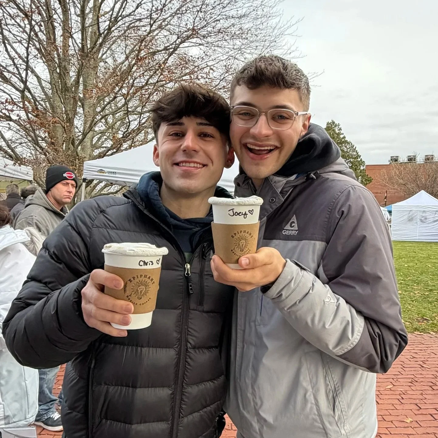 Two young men smiling and holding cups of soft serve ice cream outdoors, with a background of tents and other people, on a cloudy day.
