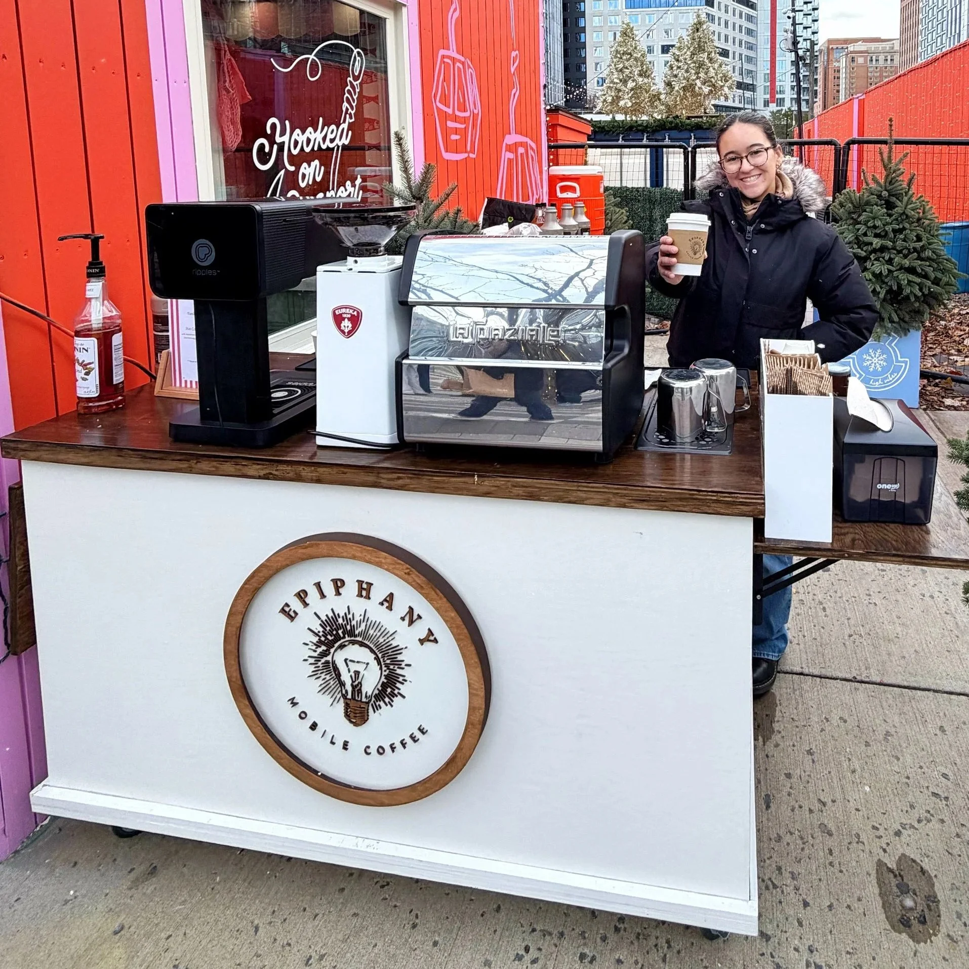 A woman standing at a mobile coffee stand with a large coffee machine, a coffee to-go cup, and a sign that says "EPI PHANY MOBILE COFFEE." She is wearing glasses, a black coat with a fur-lined hood, and smiling while holding a coffee cup outdoors in 