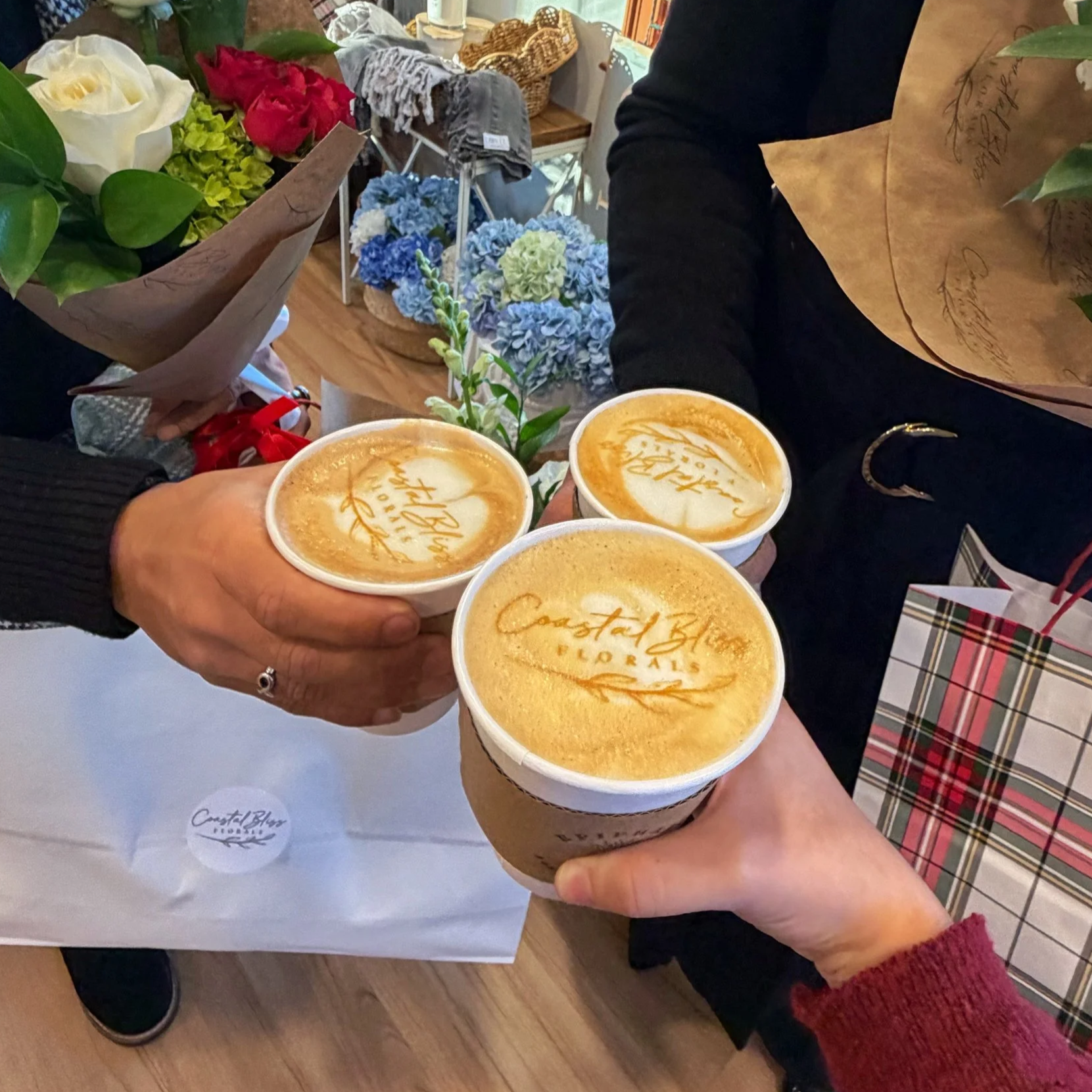 Three people holding cups of coffee with foam art that reads 'Coastal Blue Florals' and 'Happy Holidays' in a floral shop, with bouquets of flowers and gift bags nearby.