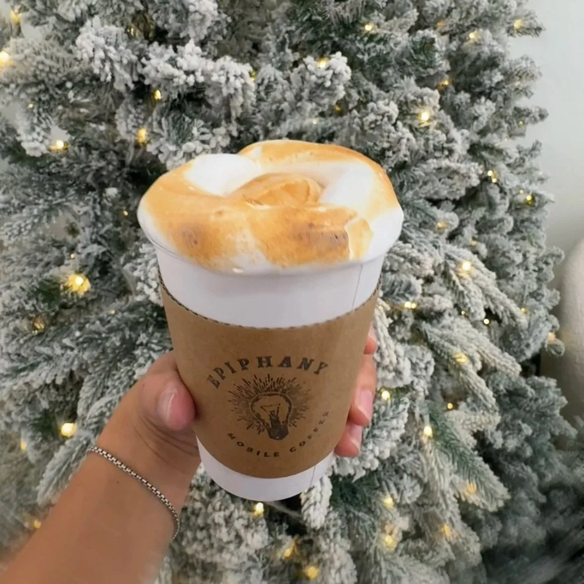 Hand holding a caramel frappuccino with whipped cream in front of a snow-flocked Christmas tree with small lights.