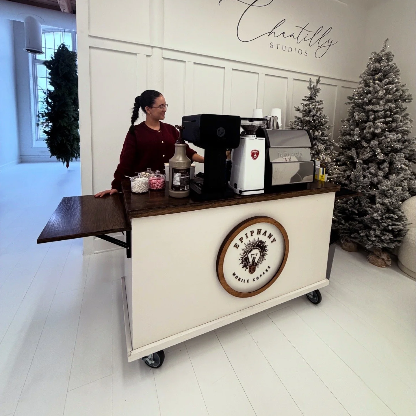 A woman standing behind a mobile coffee cart with the sign 'Piphany Mobile Coffee,' decorated with Christmas trees and holiday decor in the background.