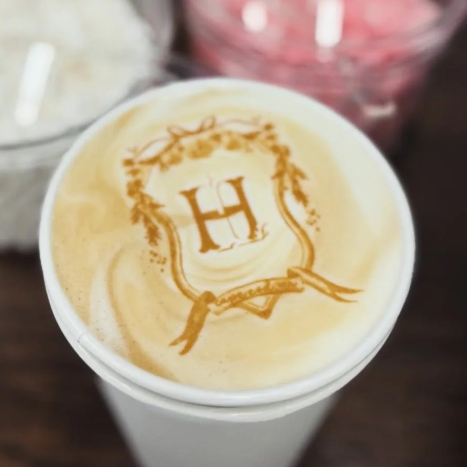 A coffee cup with a latte art design featuring a large letter H within a crest, surrounded by decorative elements.