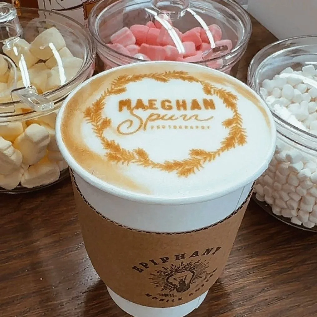 A hot beverage with latte art that reads "Maeghan" and "Spirehan" in a foam design, surrounded by bowls of colorful marshmallows in pink, yellow, and white on a wooden table.