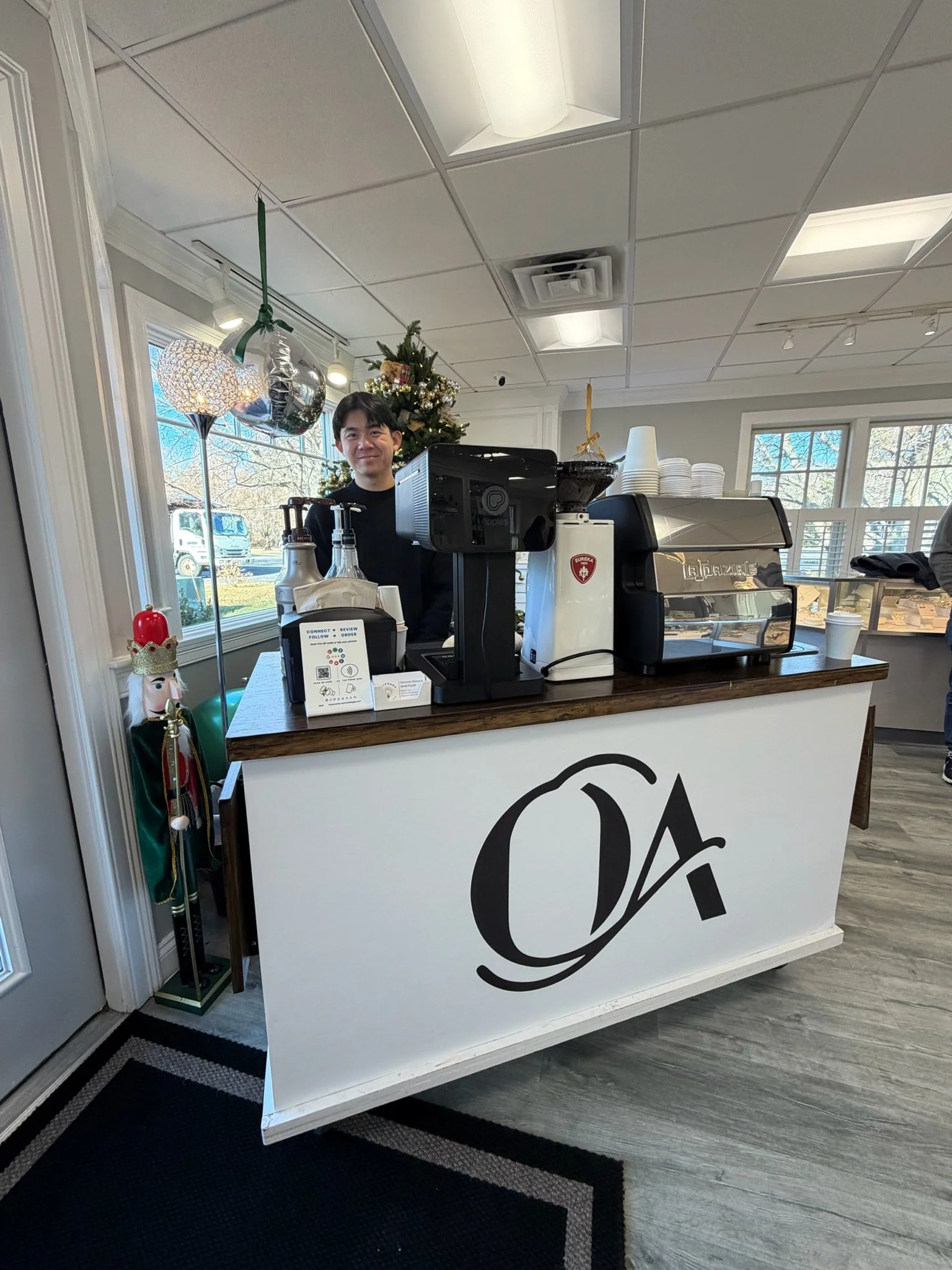 A smiling barista stands behind a coffee counter decorated with Christmas ornaments, including a small Christmas tree and balloons, inside a bright cafe with large windows.