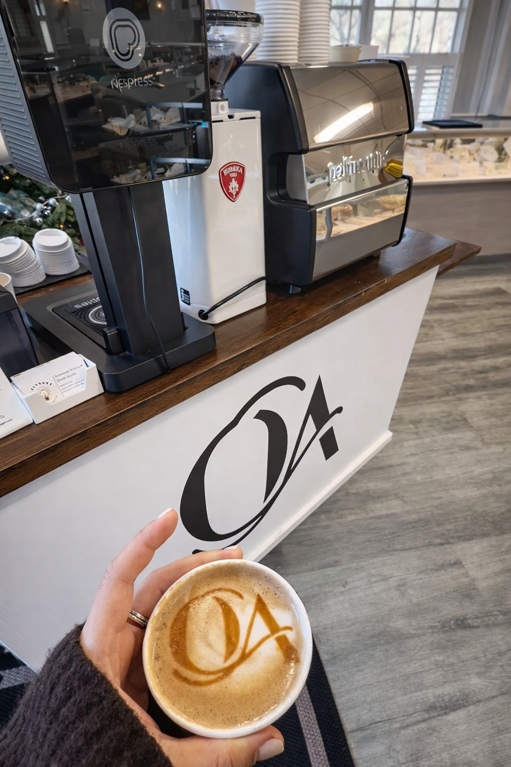Person holding a coffee cup with a logo on top, in front of a coffee station with brewing equipment and cups.