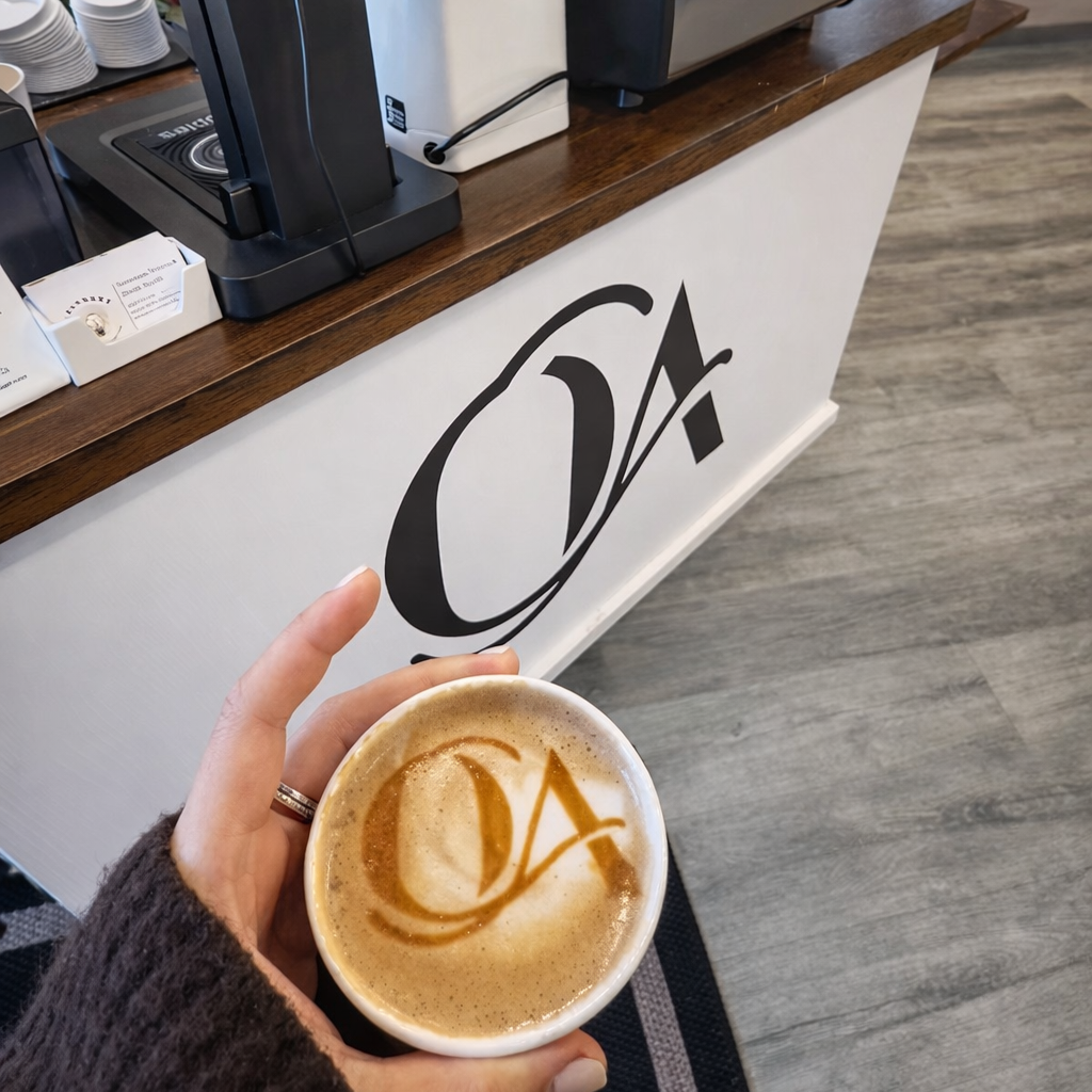 A person holding a coffee cup with latte art logo in a coffee shop. The logo on the coffee matches the logo on the counter.