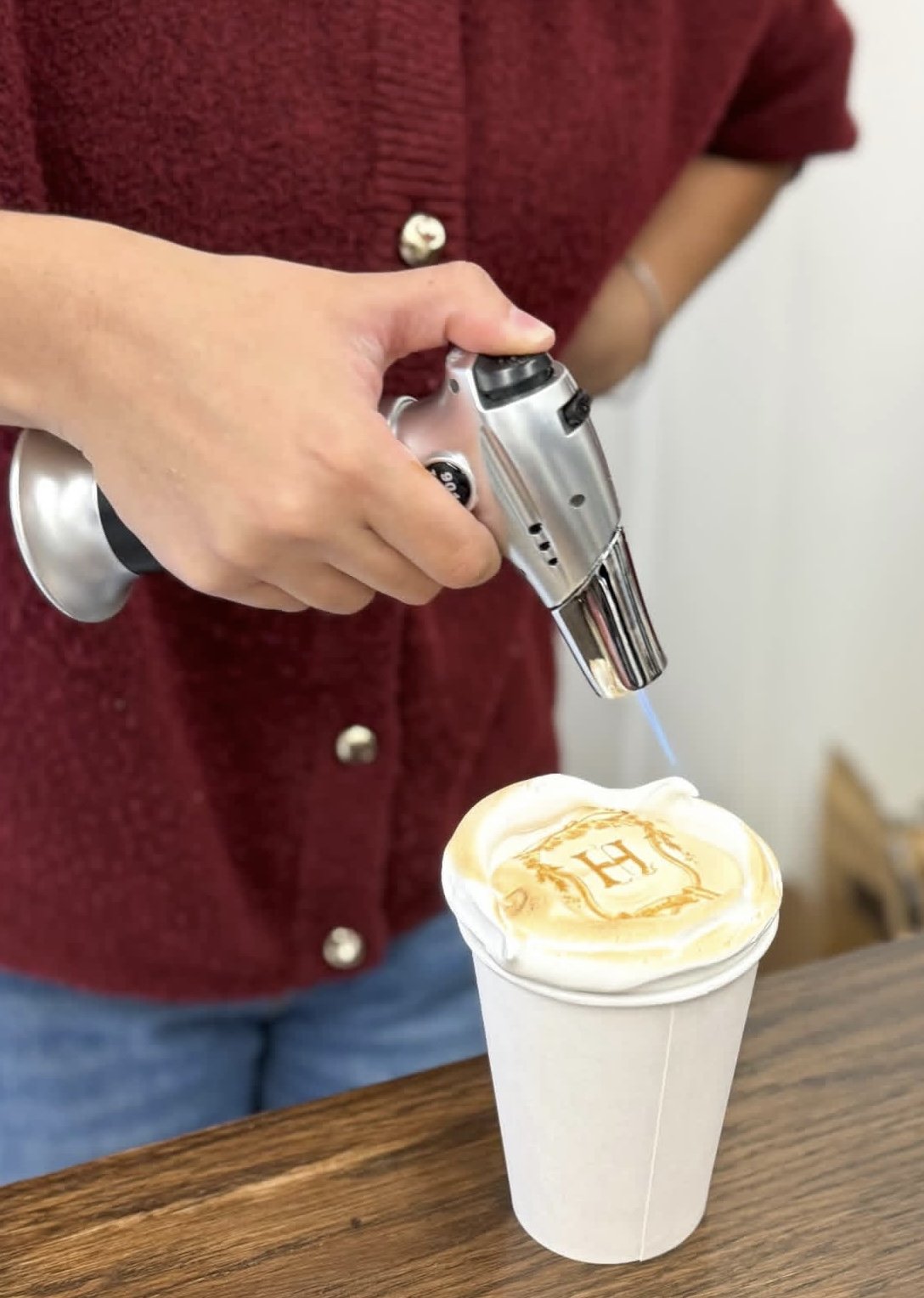 Person using a culinary torch to caramelize toppings on a dessert served in a white cup with a monogrammed design on top.