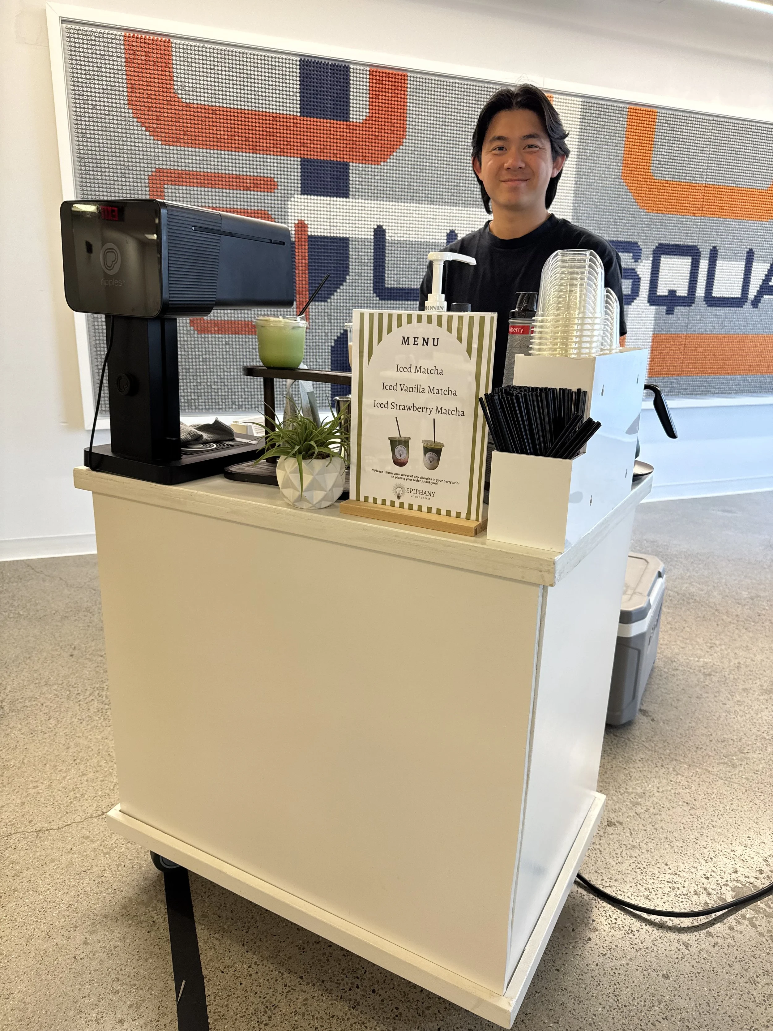 A person standing behind a white mobile cart with drink supplies, including disposable cups, a plant, and a menu sign listing flavored matcha drinks. There is a drink machine and a container of lids on the cart. The background has a decorative wall w